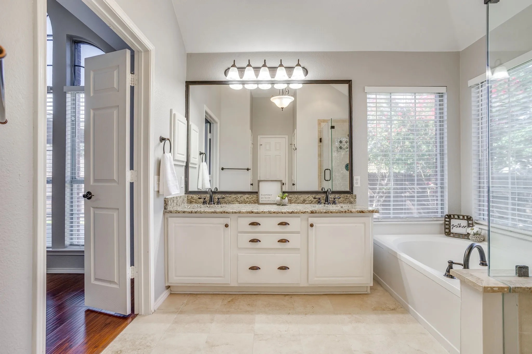 Dual vanity with a stone countertop, a built-in soaking tub, a framed mirror, and tile flooring