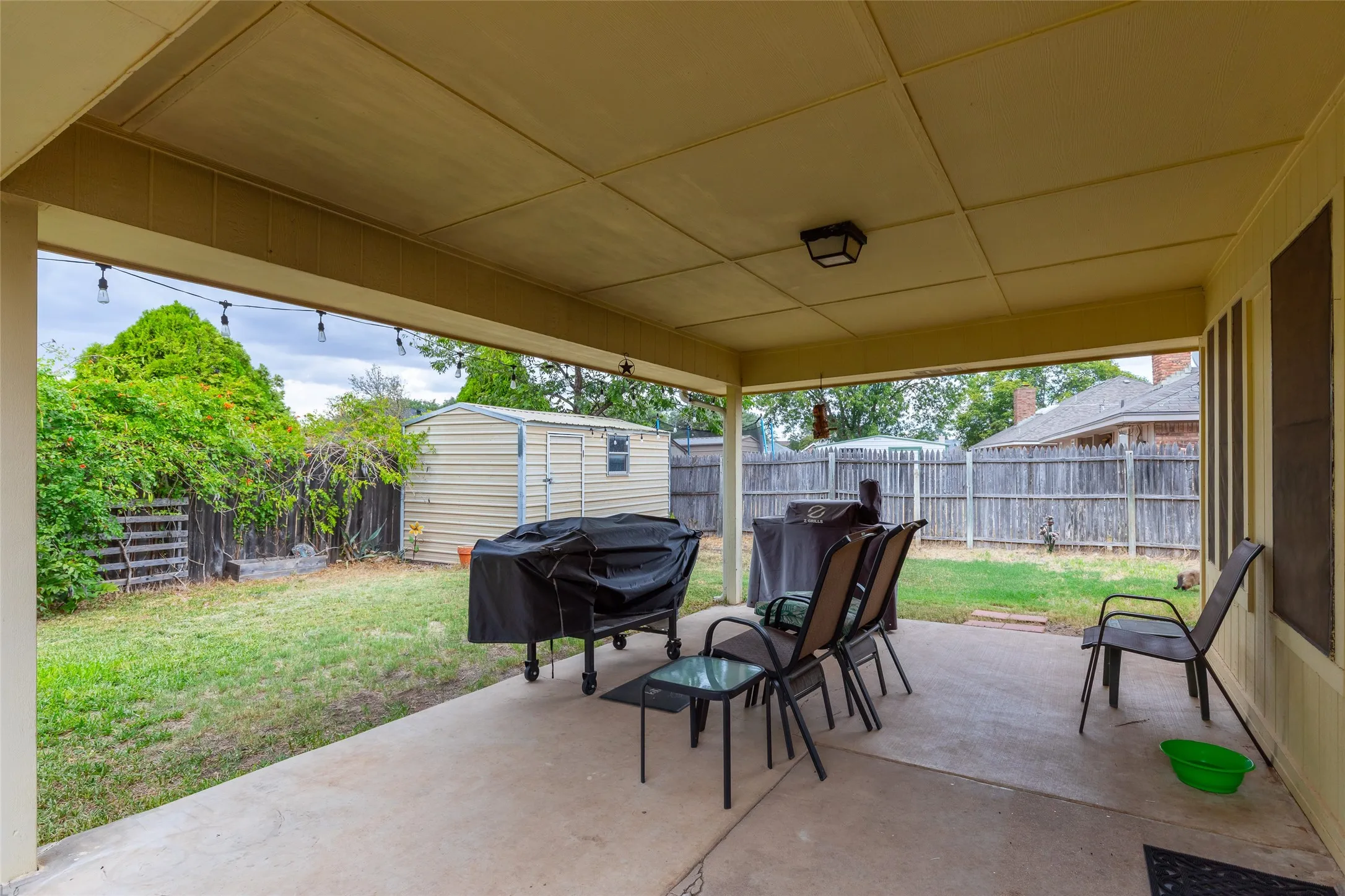 Covered concrete patio with overhead lighting, a grass lawn, and a wood privacy fence