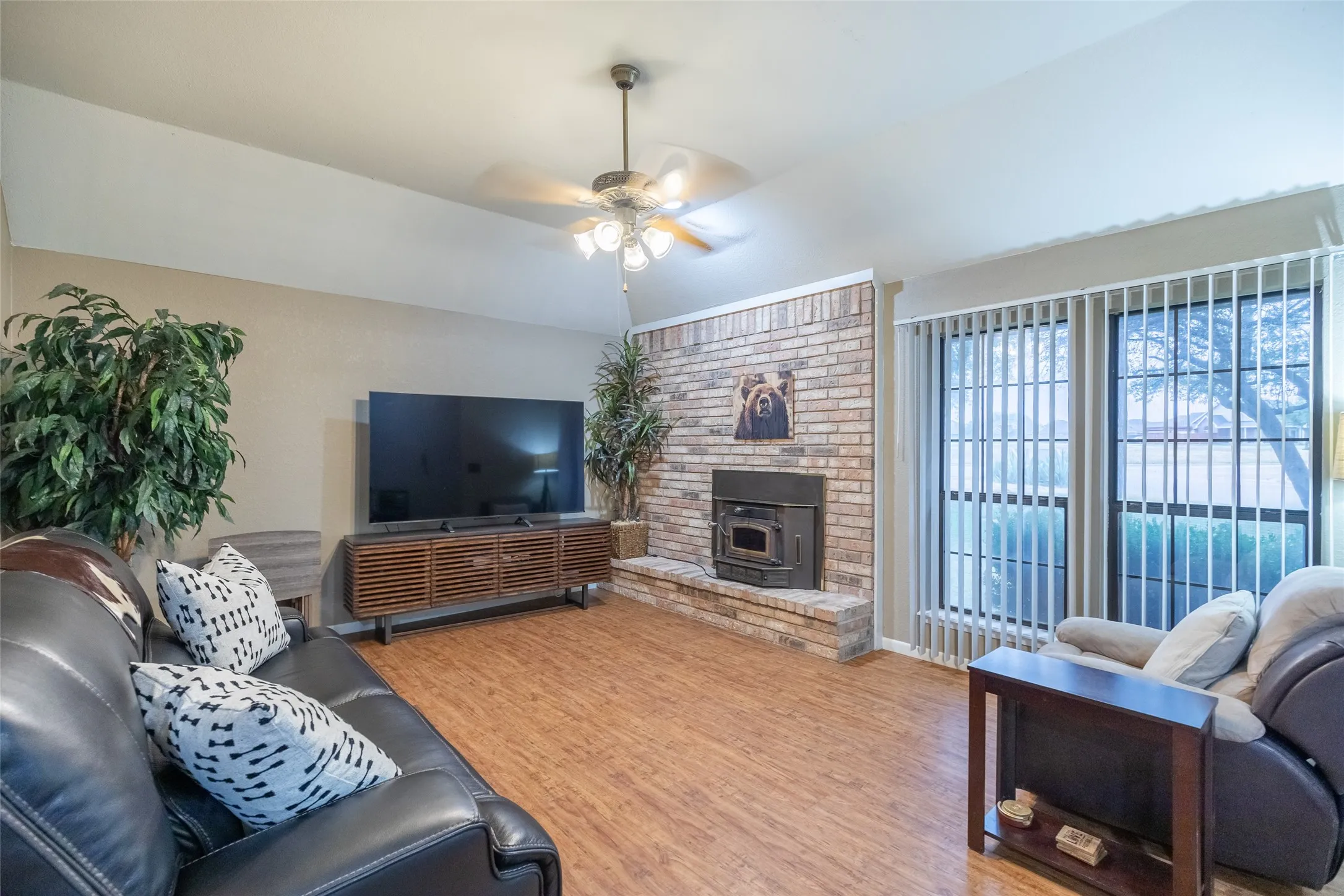 Vaulted ceiling with a ceiling fan, brick fireplace, wood-finish flooring, and vertical blinds