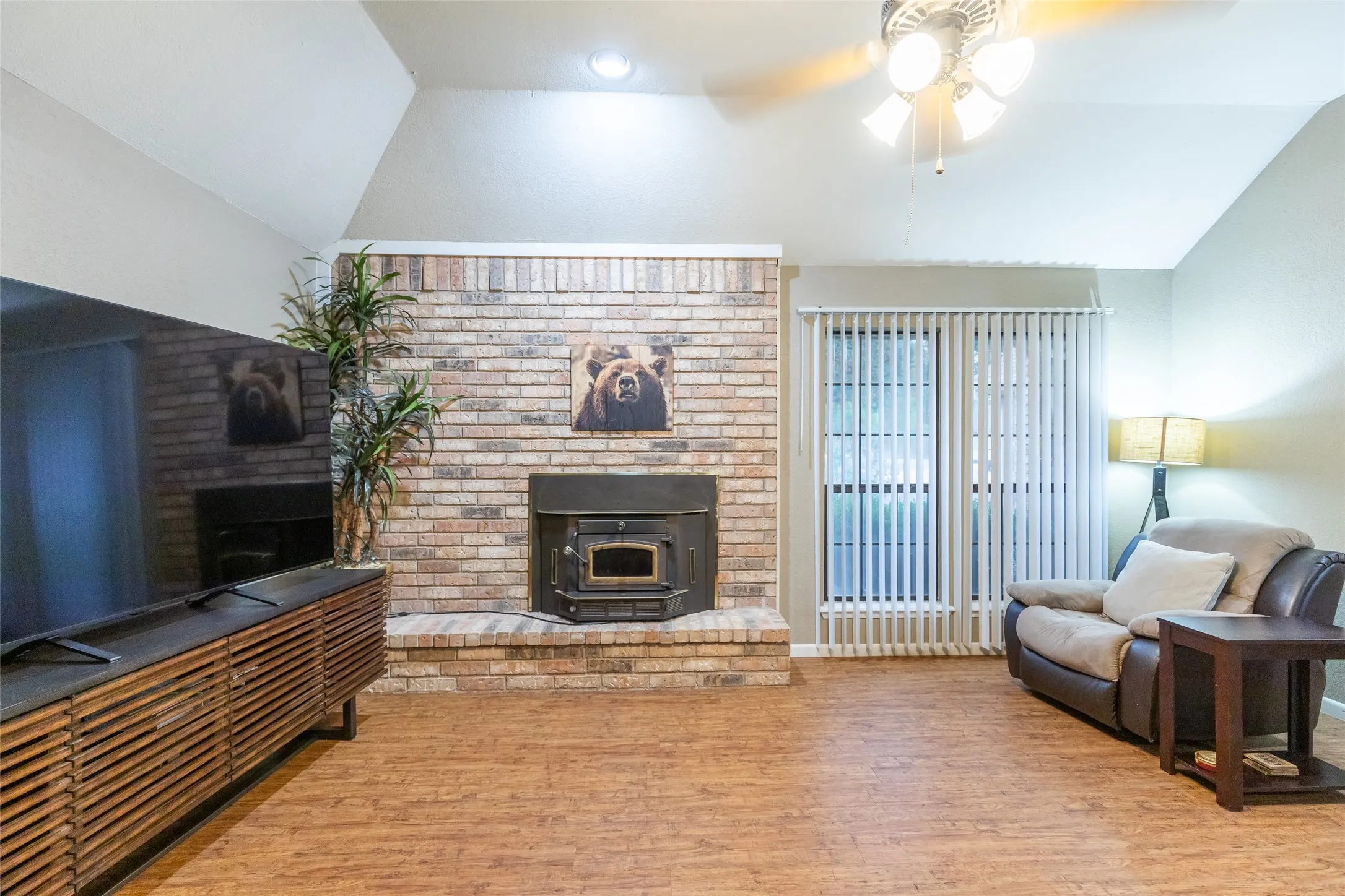 Vaulted ceiling with a ceiling fan, brick-faced fireplace with a wood-burning insert, full-light glass door with vertical blinds, and wood-finish flooring