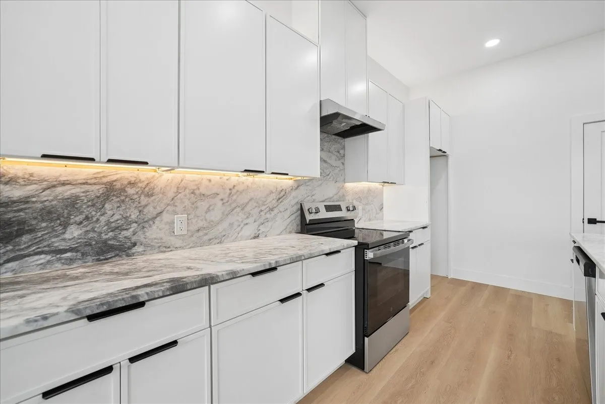 Kitchen featuring full-height white cabinetry, integrated under-cabinet lighting, natural stone countertops with matching backsplash, and wood-finish flooring