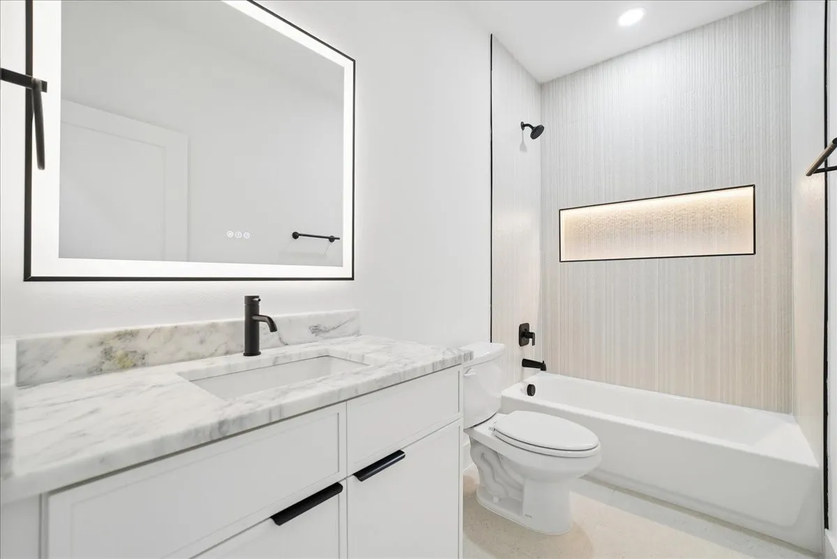 Backlit vanity mirror, single basin sink, white cabinetry with dark pull hardware, white tub, and an illuminated shower niche