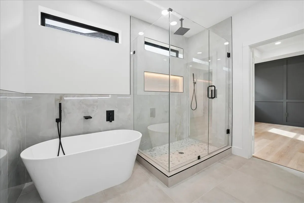 Freestanding soaking tub with matte black fixtures, glass-enclosed shower with tiled walls and rainfall showerhead, and light-toned tile flooring