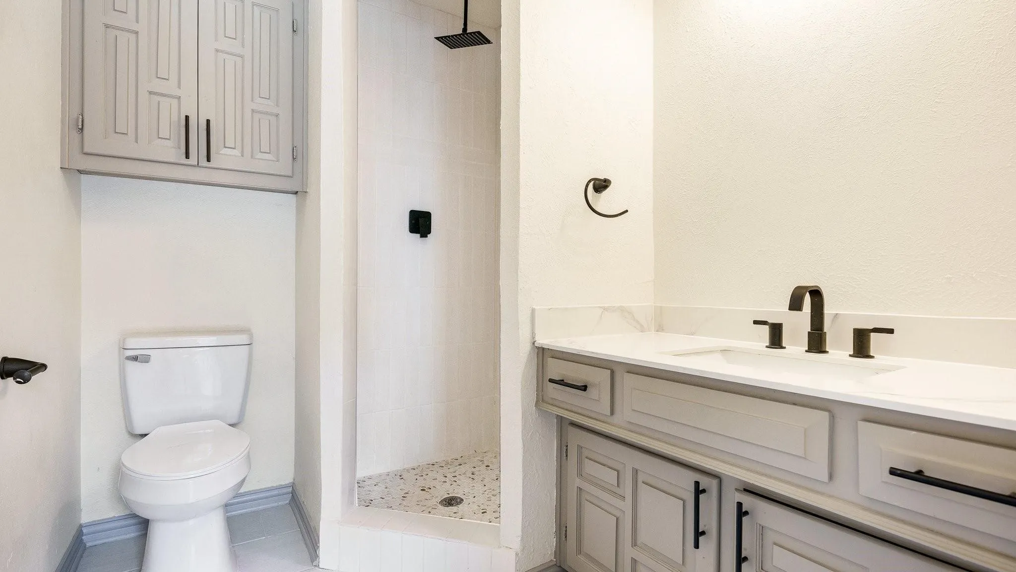 Walk-in shower with tiled walls and floor, dual vanity with light-toned cabinetry, white countertop, and dark-toned hardware, and an overhead built-in storage cabinet