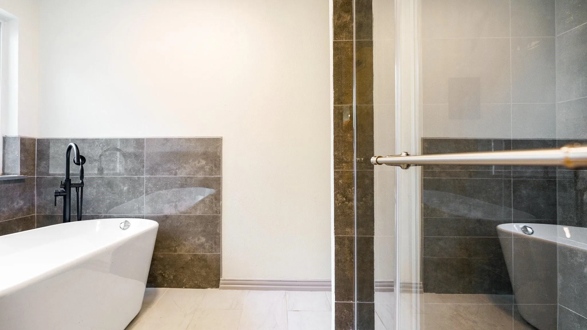 Freestanding soaking tub with a floor-mounted black faucet, glass-enclosed shower, and large format wall tiling