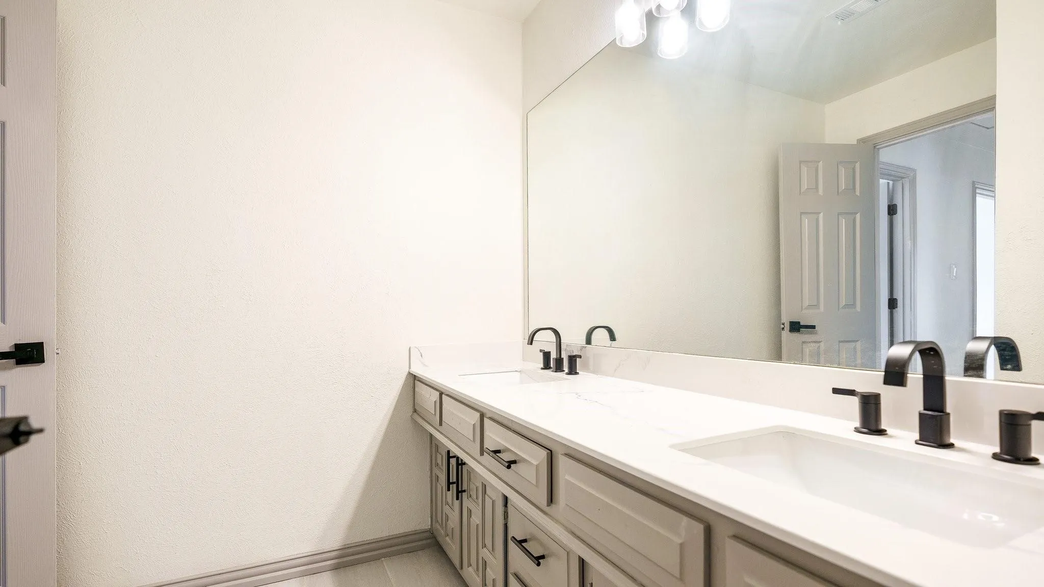 Dual undermount sinks with matte black fixtures, an expansive vanity mirror, light-toned cabinetry with dark hardware, and a stone-finish countertop
