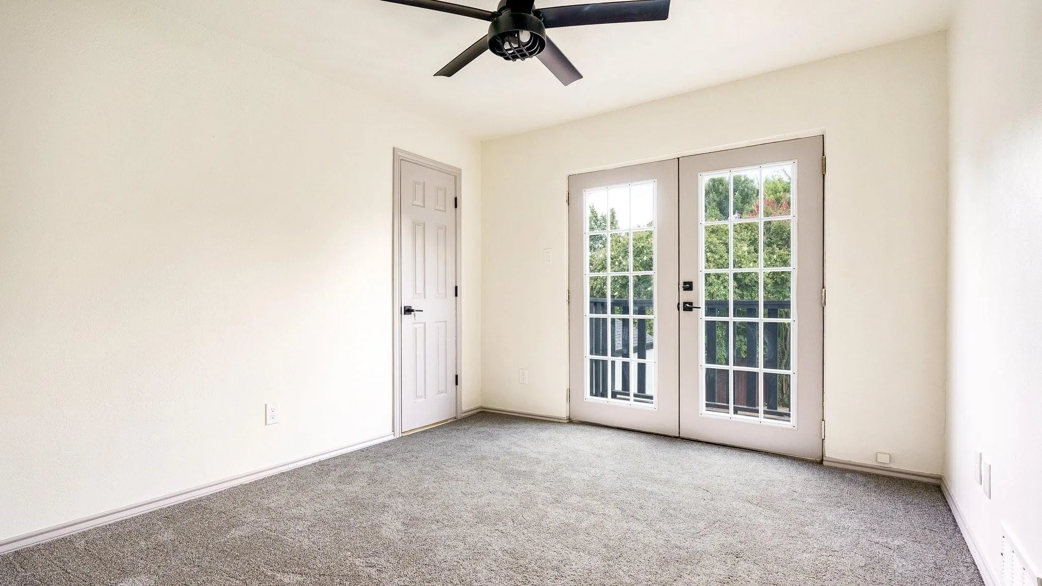 Neutral-toned wall paint, dark grey carpet flooring, black ceiling fan, white interior door with black hardware, and French doors with grid-patterned glass