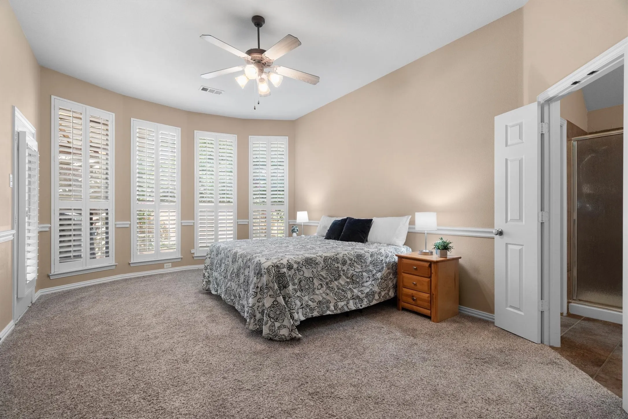 Vaulted ceiling with a ceiling fan, bay window with plantation shutters, neutral carpeting, and an attached bathroom with a shower enclosure