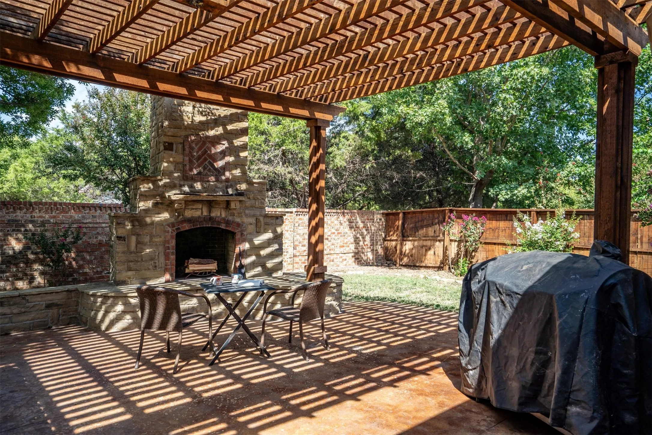 Wood pergola with slatted roof, stone outdoor fireplace, and concrete patio