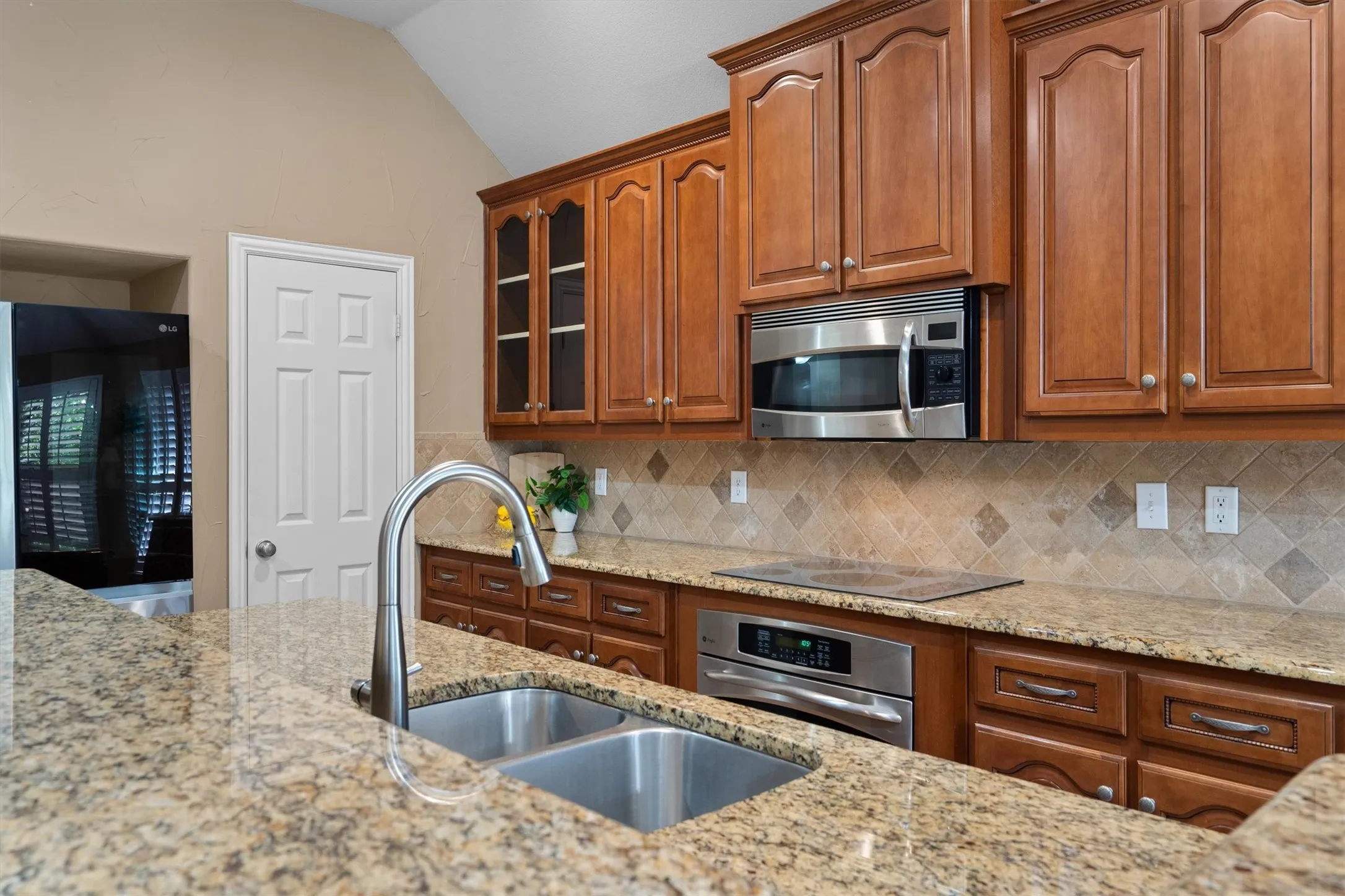 Kitchen featuring a double basin stainless steel sink, gooseneck faucet, light-toned granite countertops, wood cabinetry with glass-front accents, and stainless steel appliances including a built-in oven and microwave