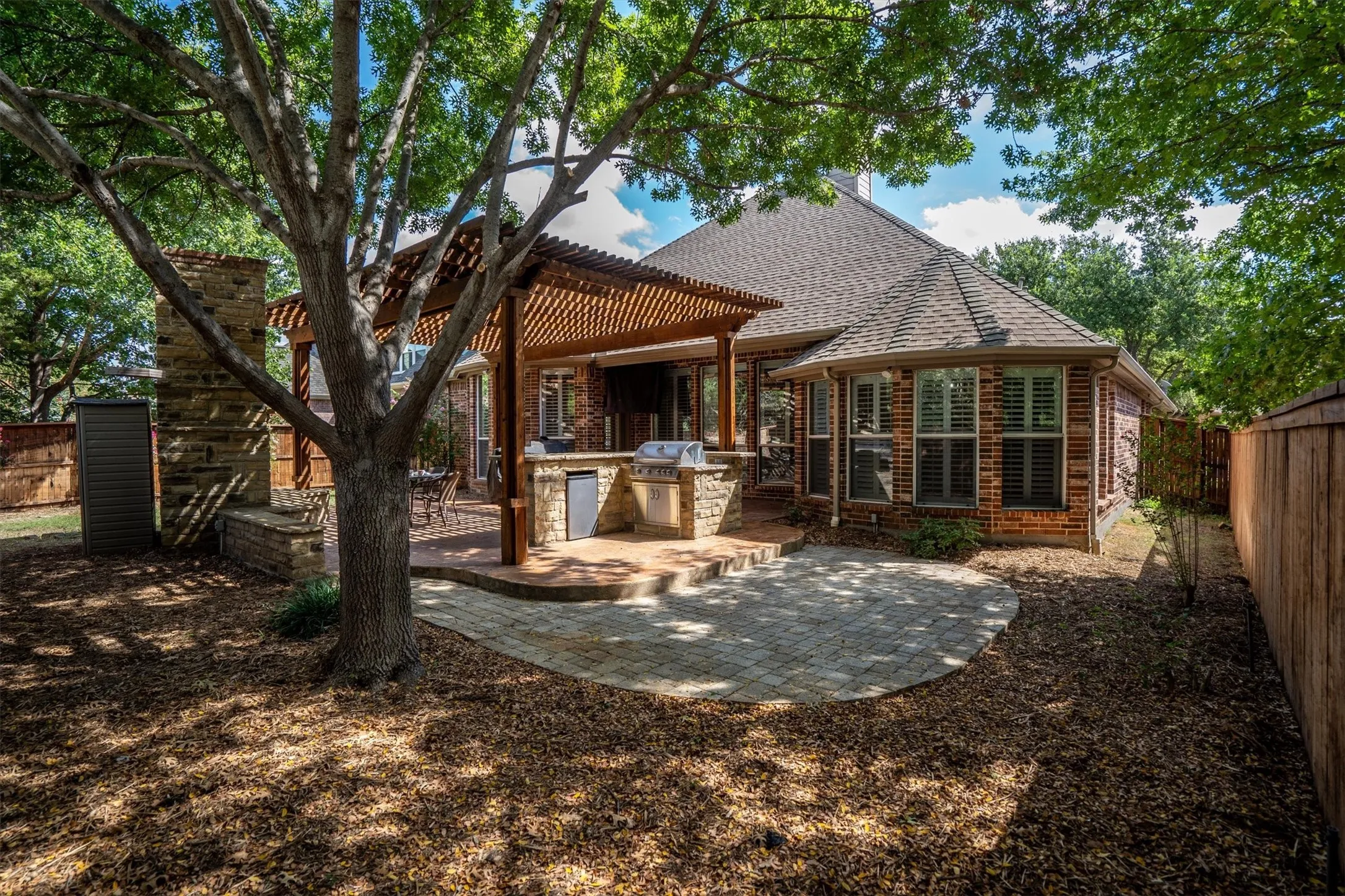 Expansive backyard featuring a stone fireplace, built-in outdoor kitchen, pergola-covered patio, and a paver patio with a curved edge