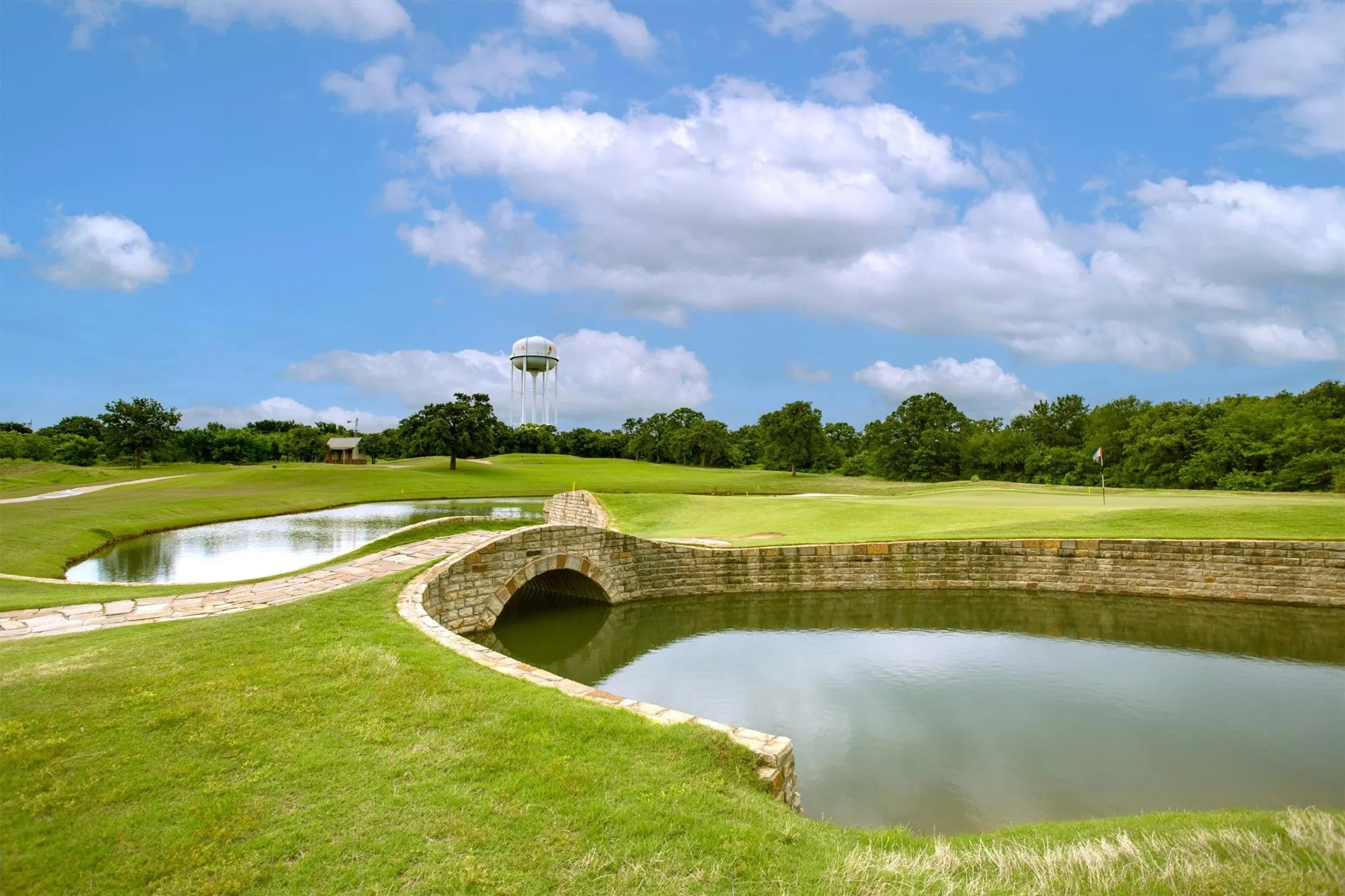 Stone arch bridge over a pond, bordered by a golf course with a visible flag, and a tree line in the distance