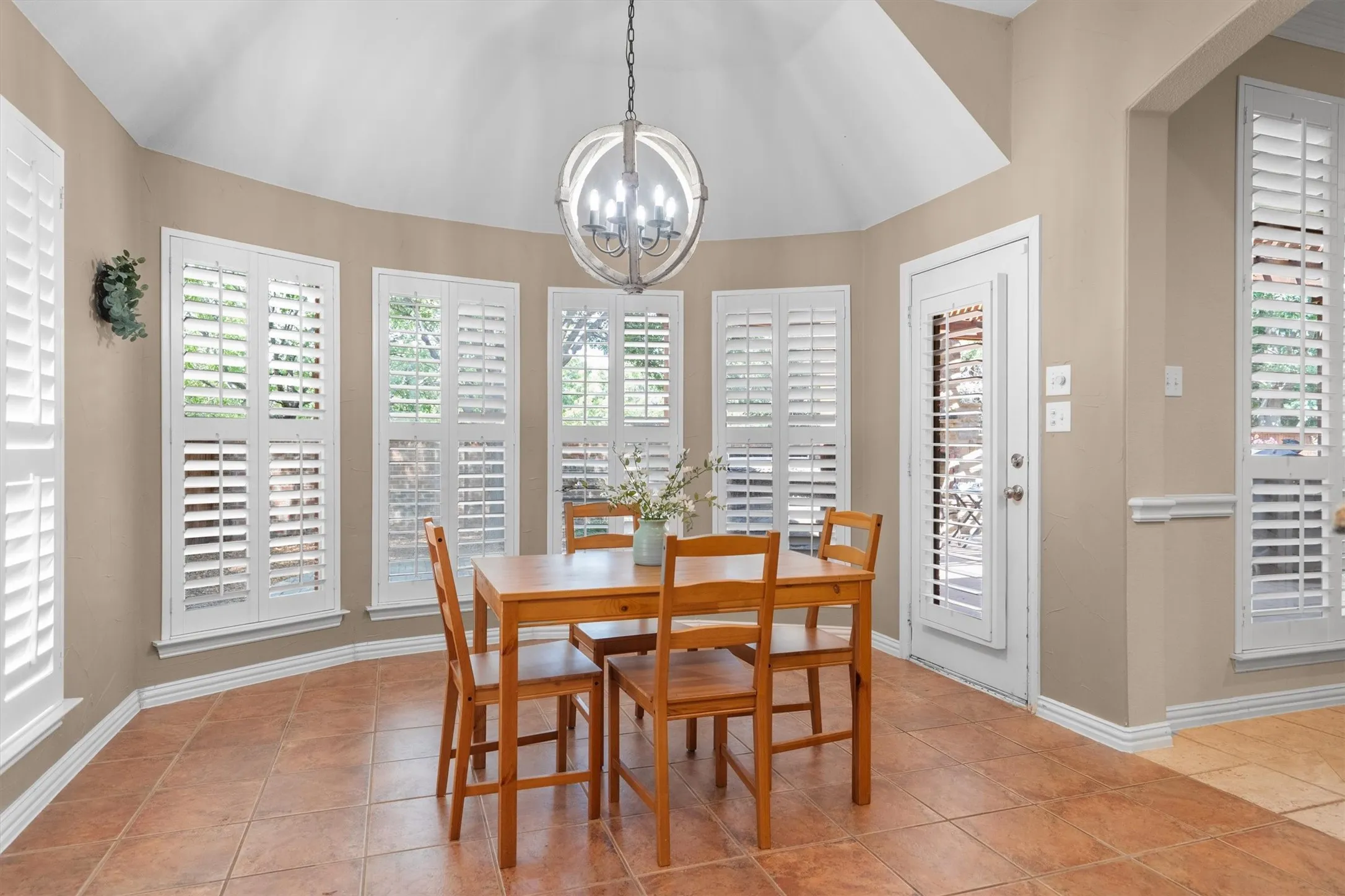 Vaulted ceiling with an orb chandelier, multiple windows with white plantation shutters, glass-paneled exterior door, and tile flooring