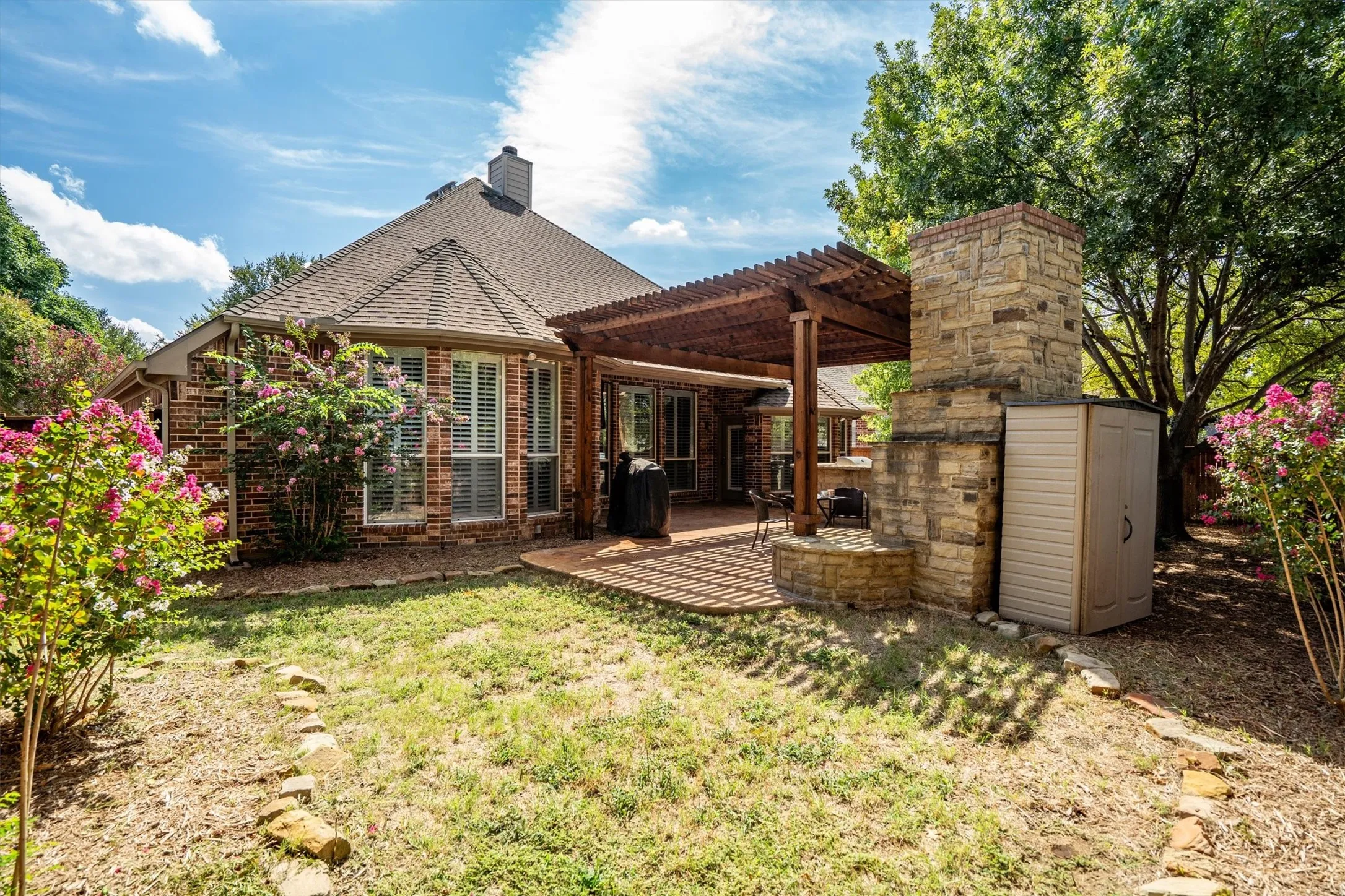 Brick exterior with multiple windows featuring plantation shutters, covered patio with wood pergola, and stone outdoor fireplace
