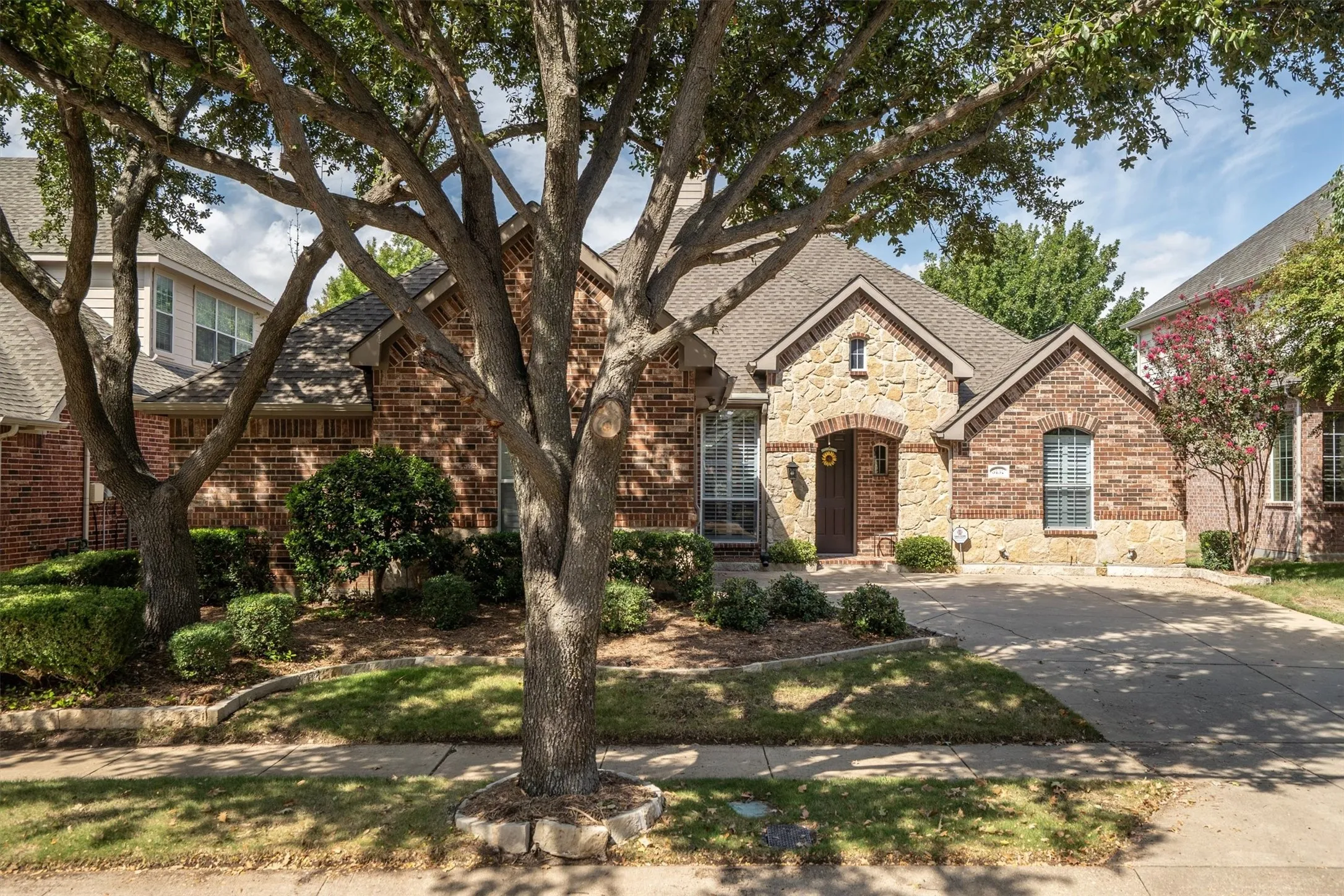 Brick and stone facade with arched entry, mature trees, and a concrete driveway