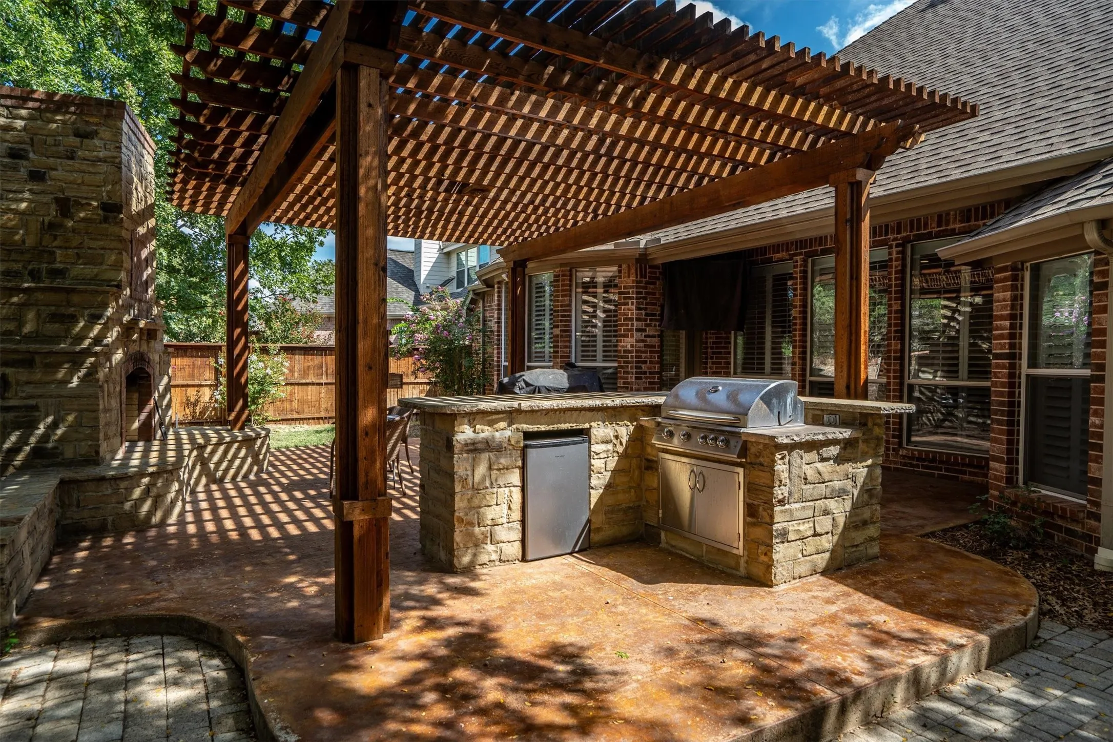 Expansive outdoor patio with a wooden pergola, built-in stone grill station, integrated outdoor refrigerator, and a stone outdoor fireplace
