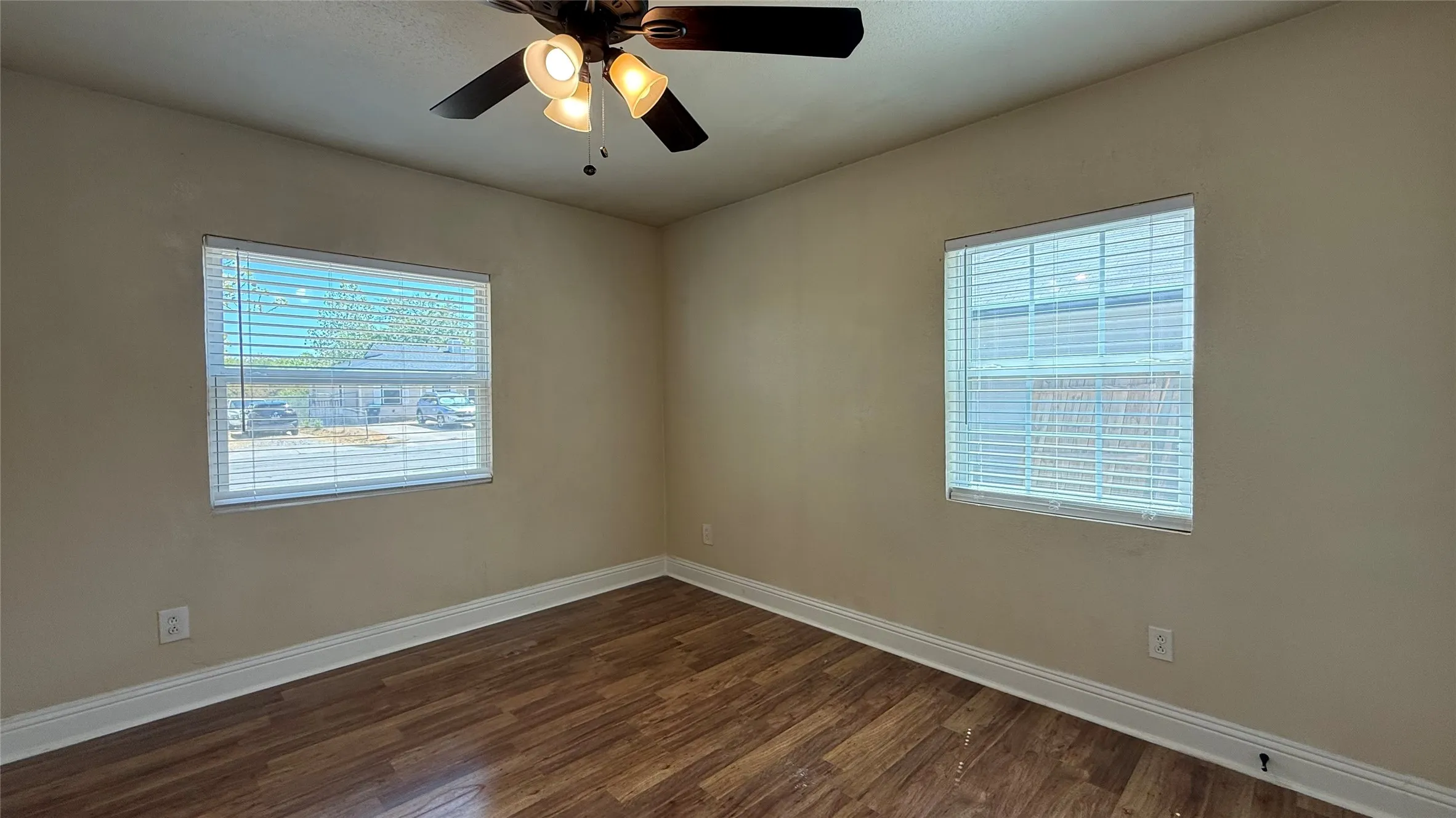 Wood-finish flooring with white baseboards, windows with blinds, and an attached ceiling fan with lighting