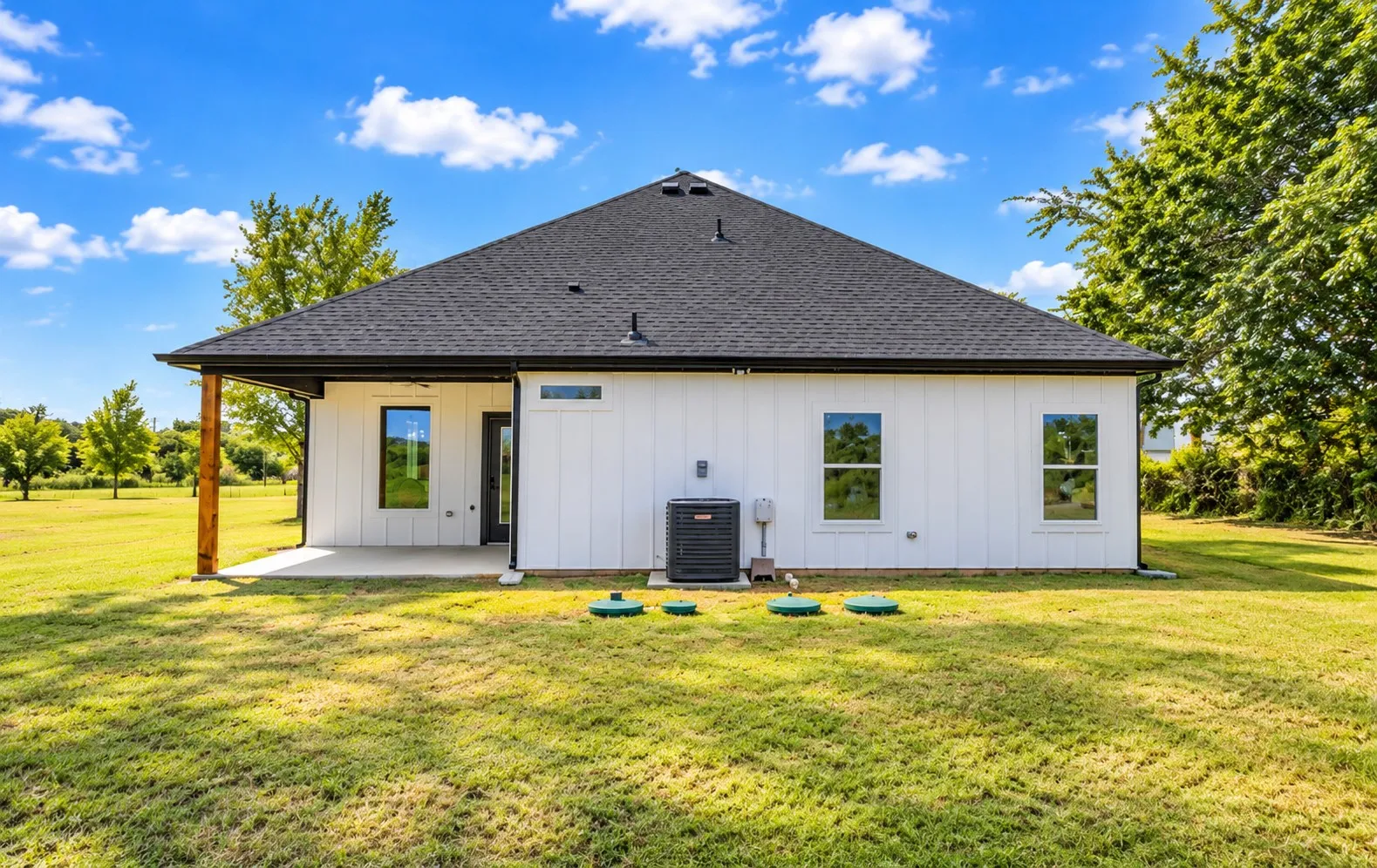 White vertical siding with black-framed windows, a dark shingle roof, and a covered concrete patio featuring a wood support beam