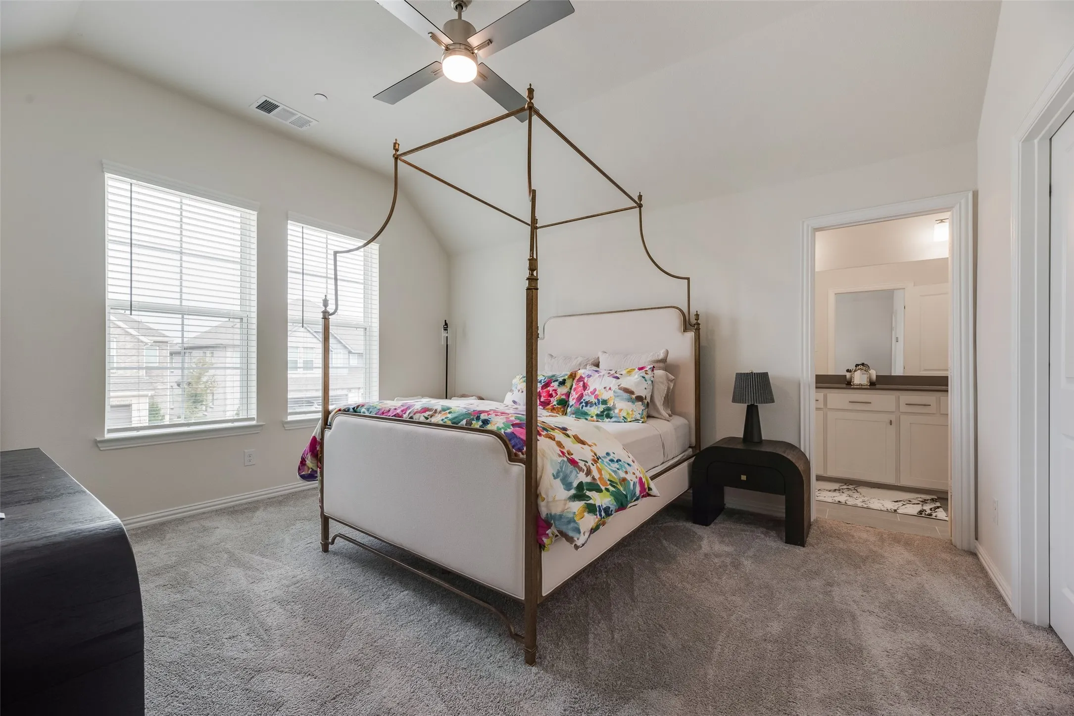 Vaulted ceiling, ceiling fan, two windows with blinds, carpeted flooring, and an adjoining bathroom entry