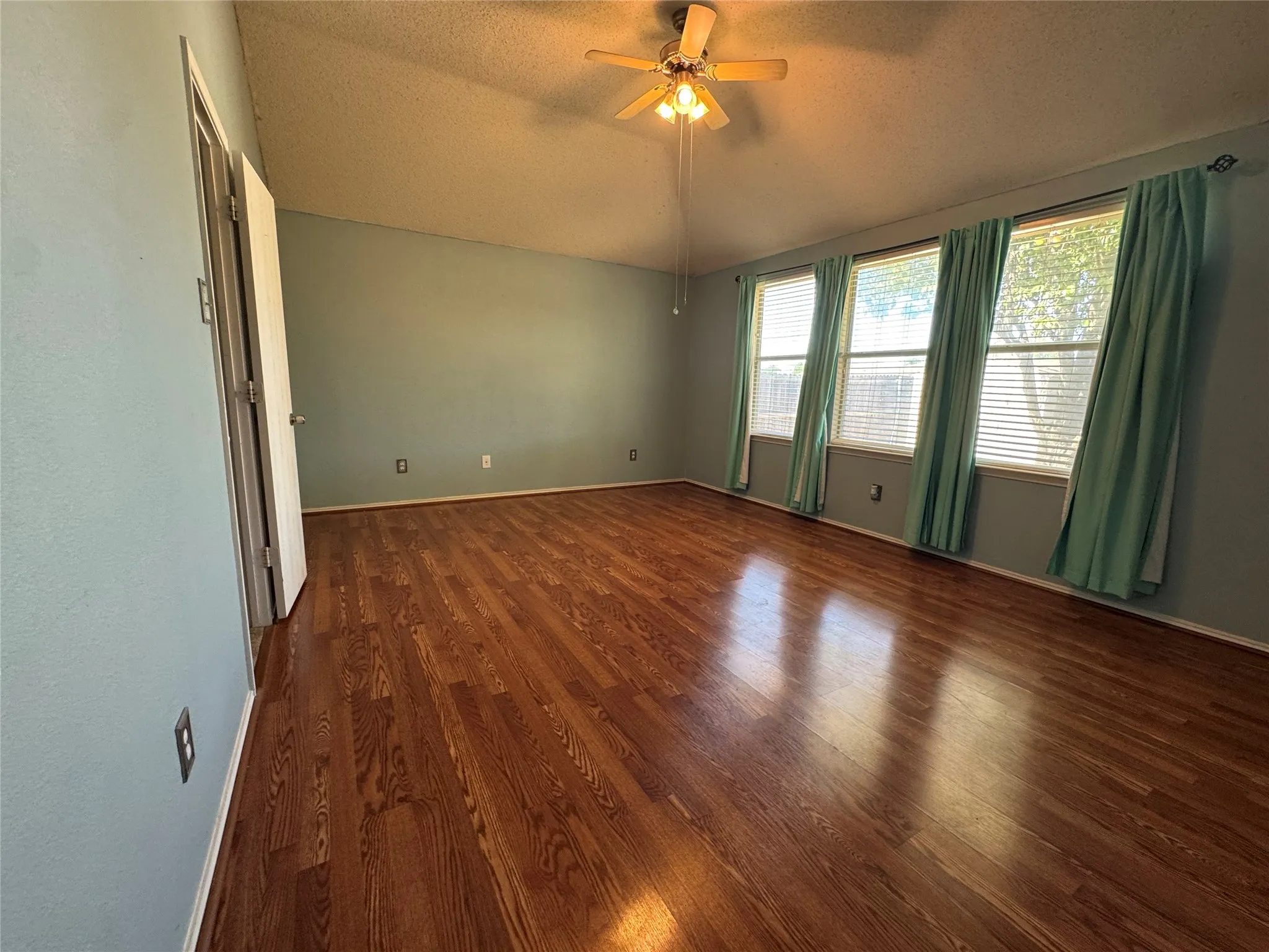 Wood-finish flooring, vaulted ceiling, ceiling fan with light fixture, three windows with blinds, and light blue walls