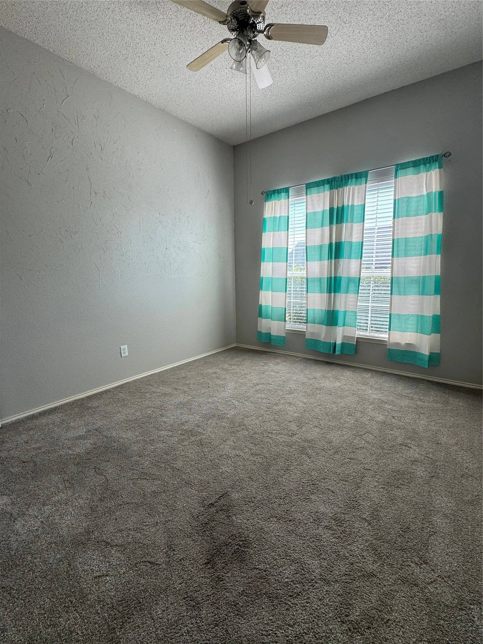 Gray textured walls with a contrasting white ceiling, ceiling fan with light fixture, window with blinds, and gray carpeted flooring