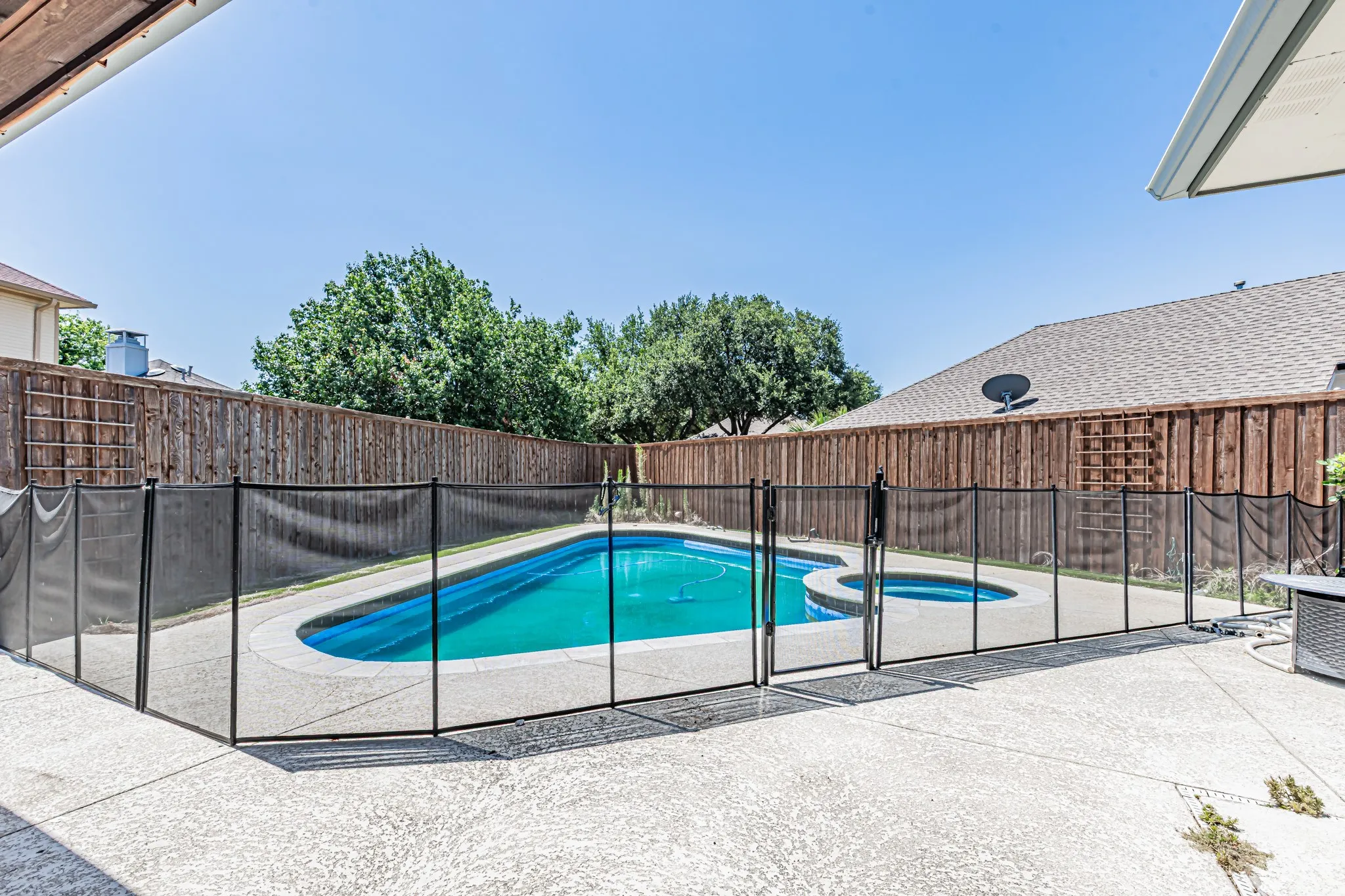 In-ground swimming pool with an integrated spa, surrounded by a mesh safety fence, wood privacy fencing, and a concrete patio