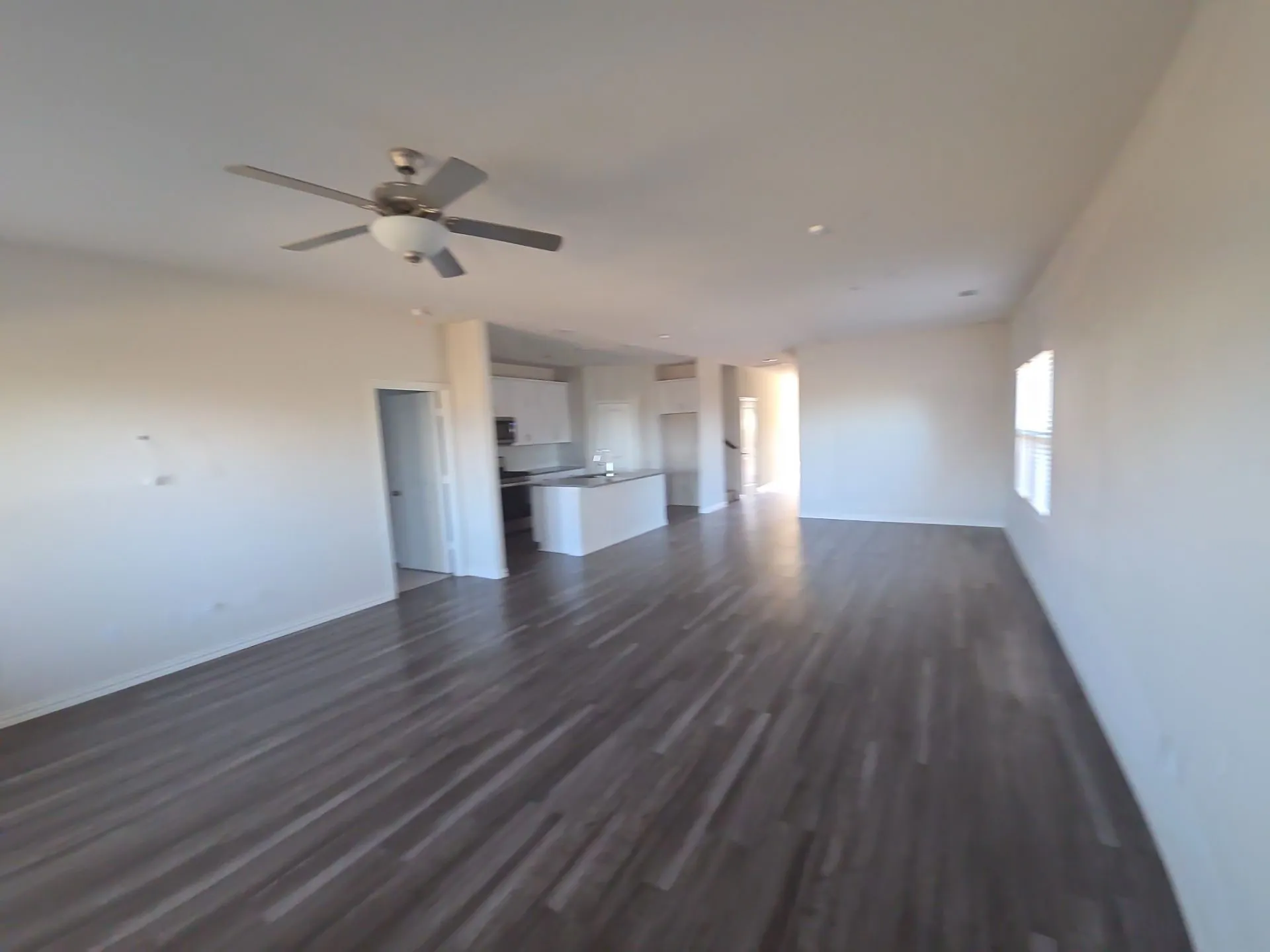 Open-concept living area featuring wood-finish flooring, a ceiling fan with integrated lighting, and a modern kitchen with white cabinetry