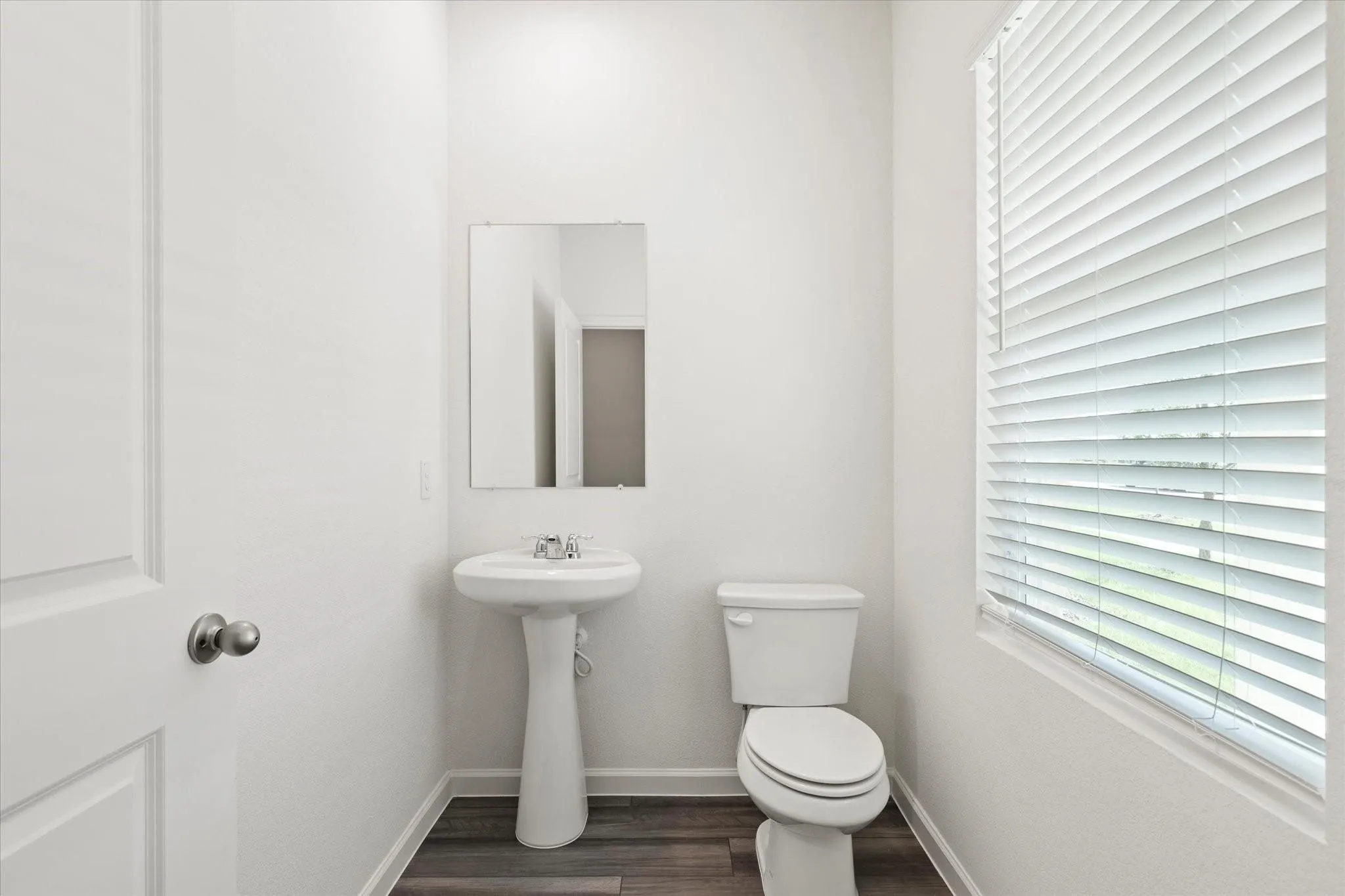 Guest Bathroom Pedestal sink with chrome faucet, wall-mounted mirror, white toilet, wood-finish flooring, and window with blinds
