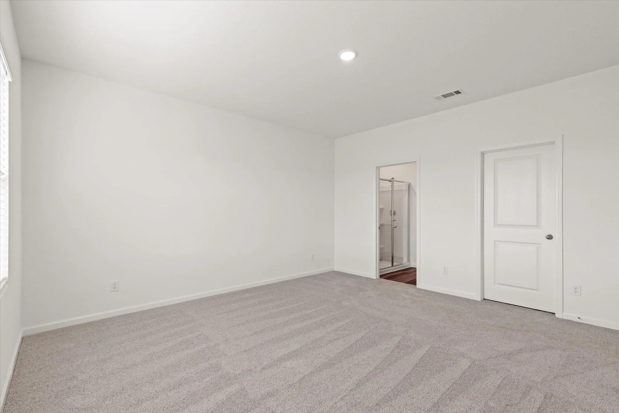 Recessed lighting, light gray carpet flooring, white painted walls, white two-panel interior door, and an adjacent bathroom with a glass-enclosed shower