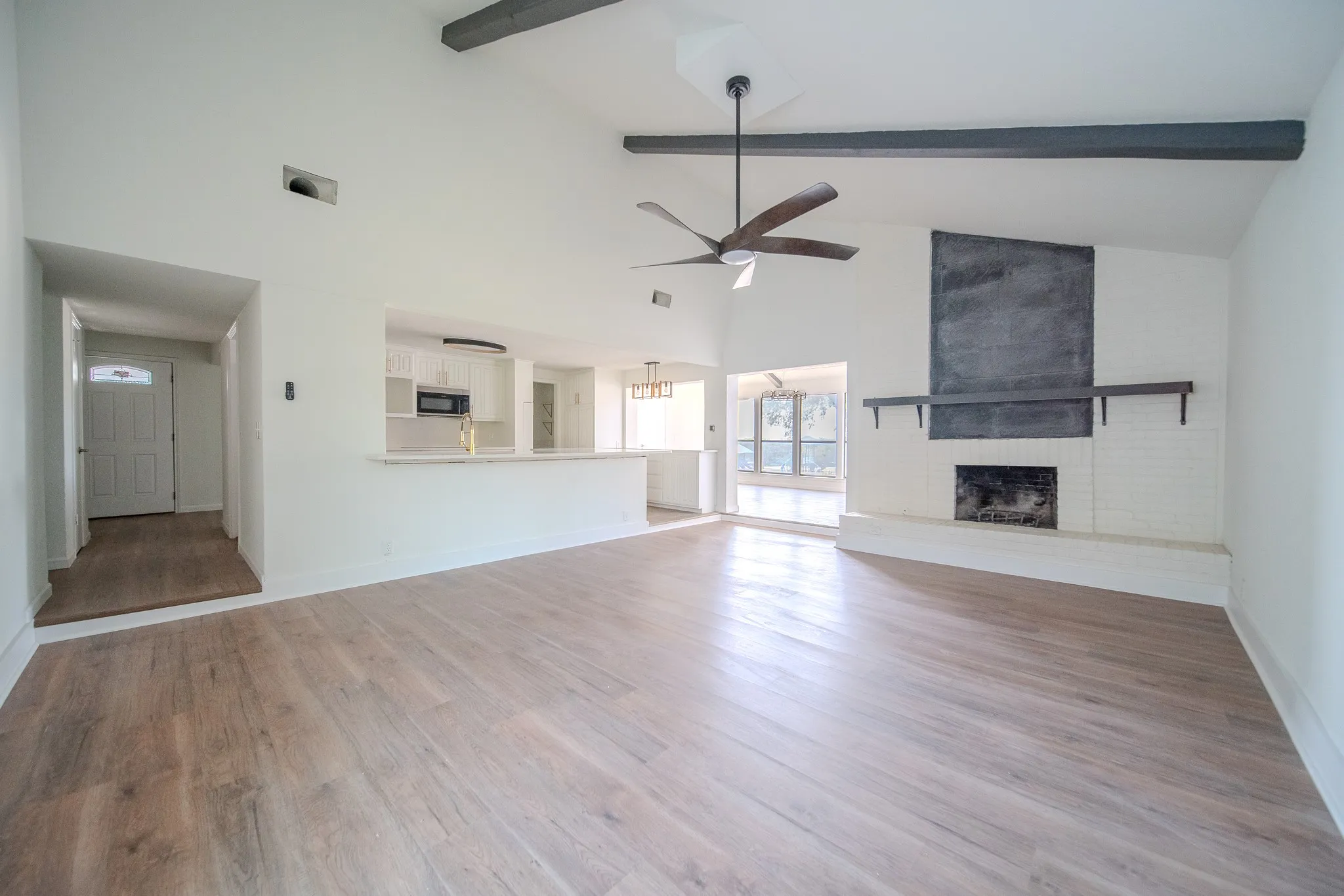 Vaulted ceiling with exposed beams, wood-finish flooring, white brick fireplace with dark mantle, ceiling fan, and a kitchen pass-through with countertop