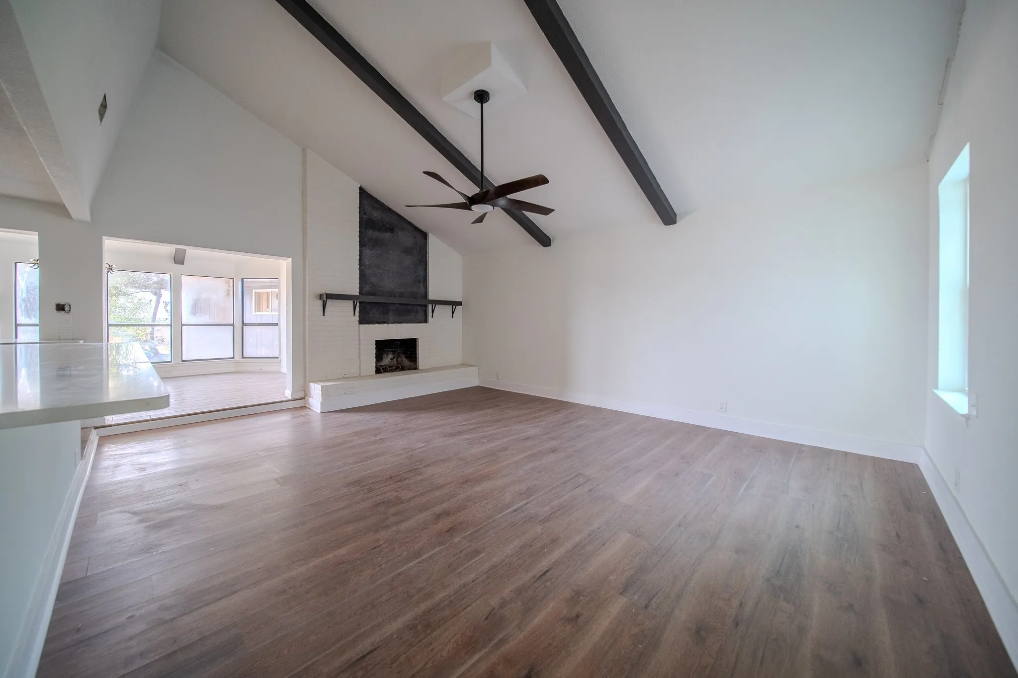 Wood-finish flooring, vaulted white ceilings with dark exposed beams, white brick fireplace with dark mantel, ceiling fan, and a built-in countertop with a light-colored surface