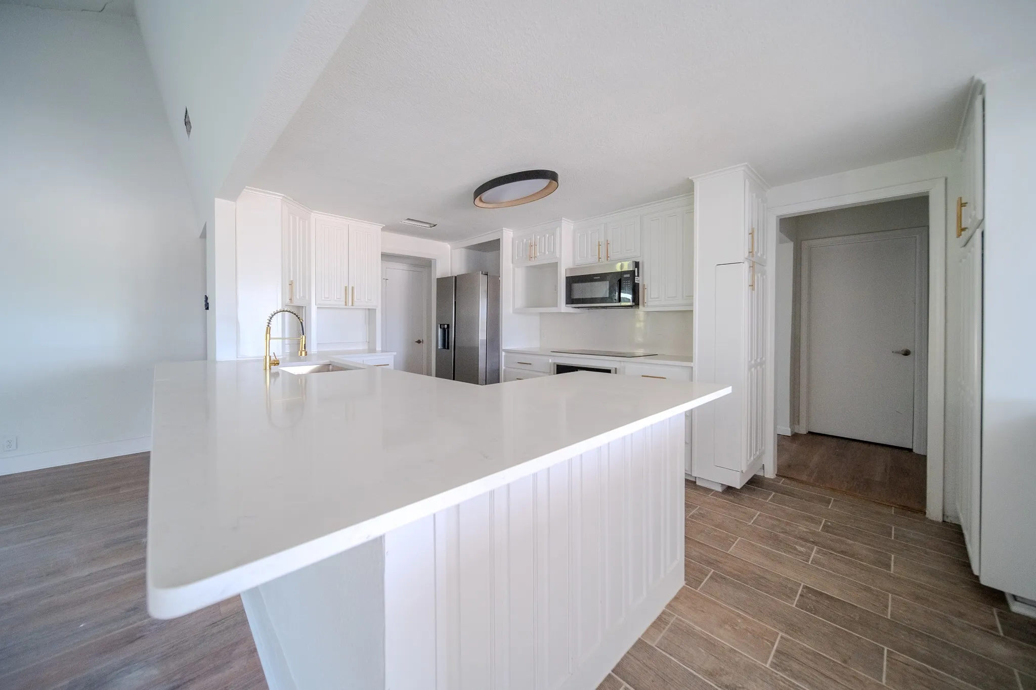 Kitchen featuring a large island with white countertop and integrated sink, white cabinetry with gold-tone hardware, stainless steel appliances, and wood-finish tile flooring