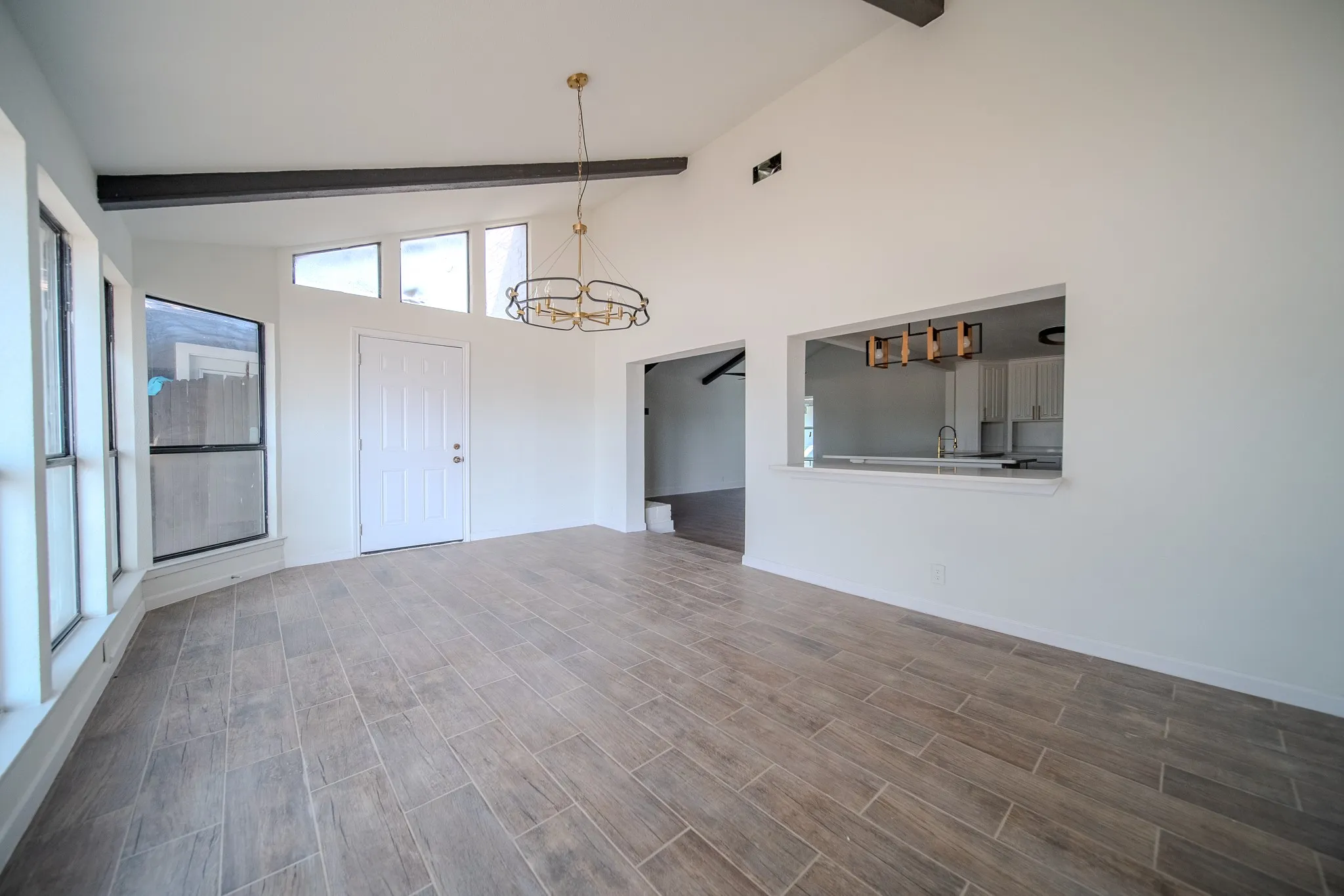Vaulted ceiling with exposed beams, wood-finish tile flooring, contemporary chandelier, large picture windows, and a pass-through window with a countertop