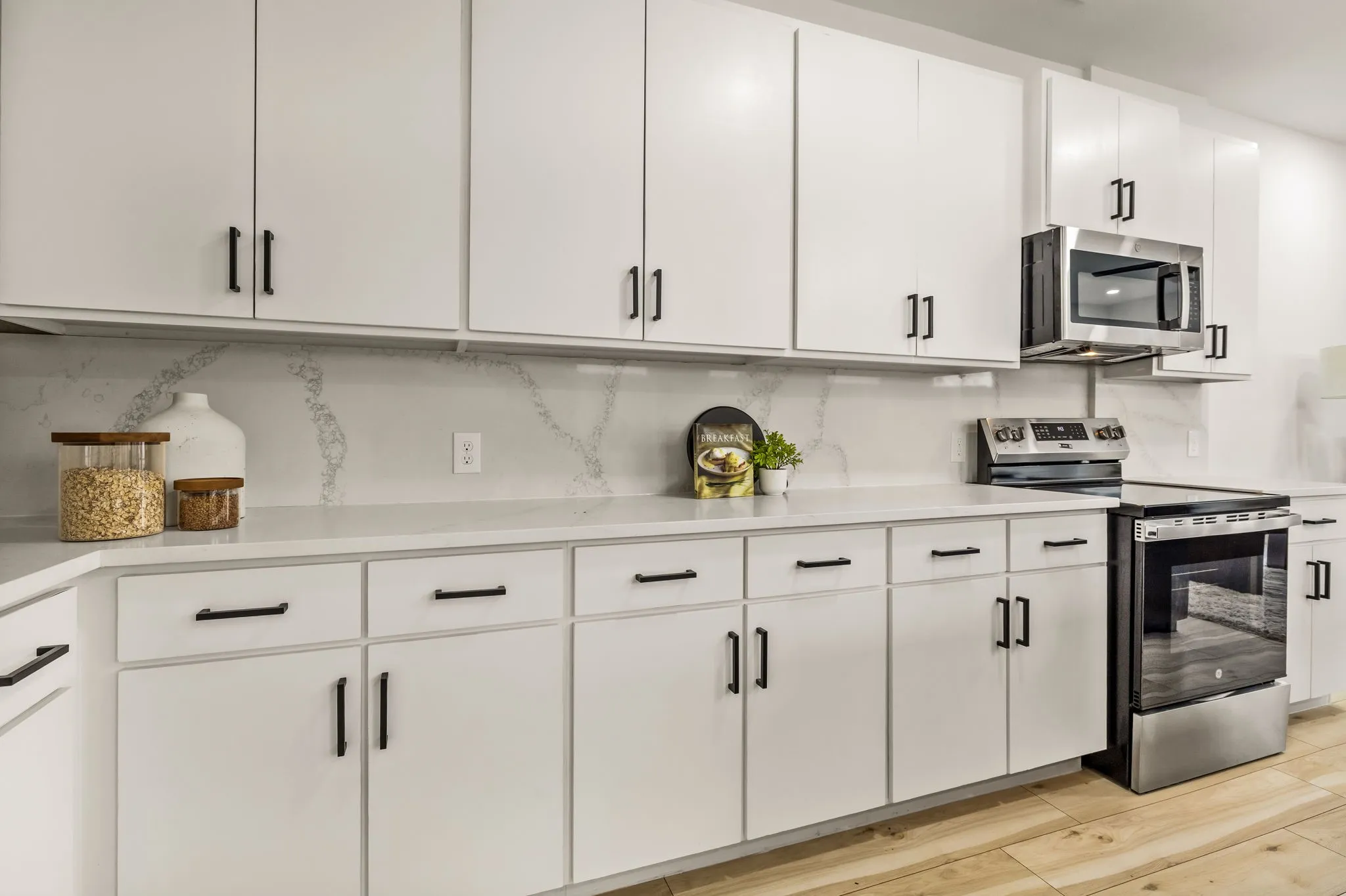 White cabinetry with matte black hardware, stainless steel appliances, light-toned wood-finish flooring, and a stone-patterned backsplash