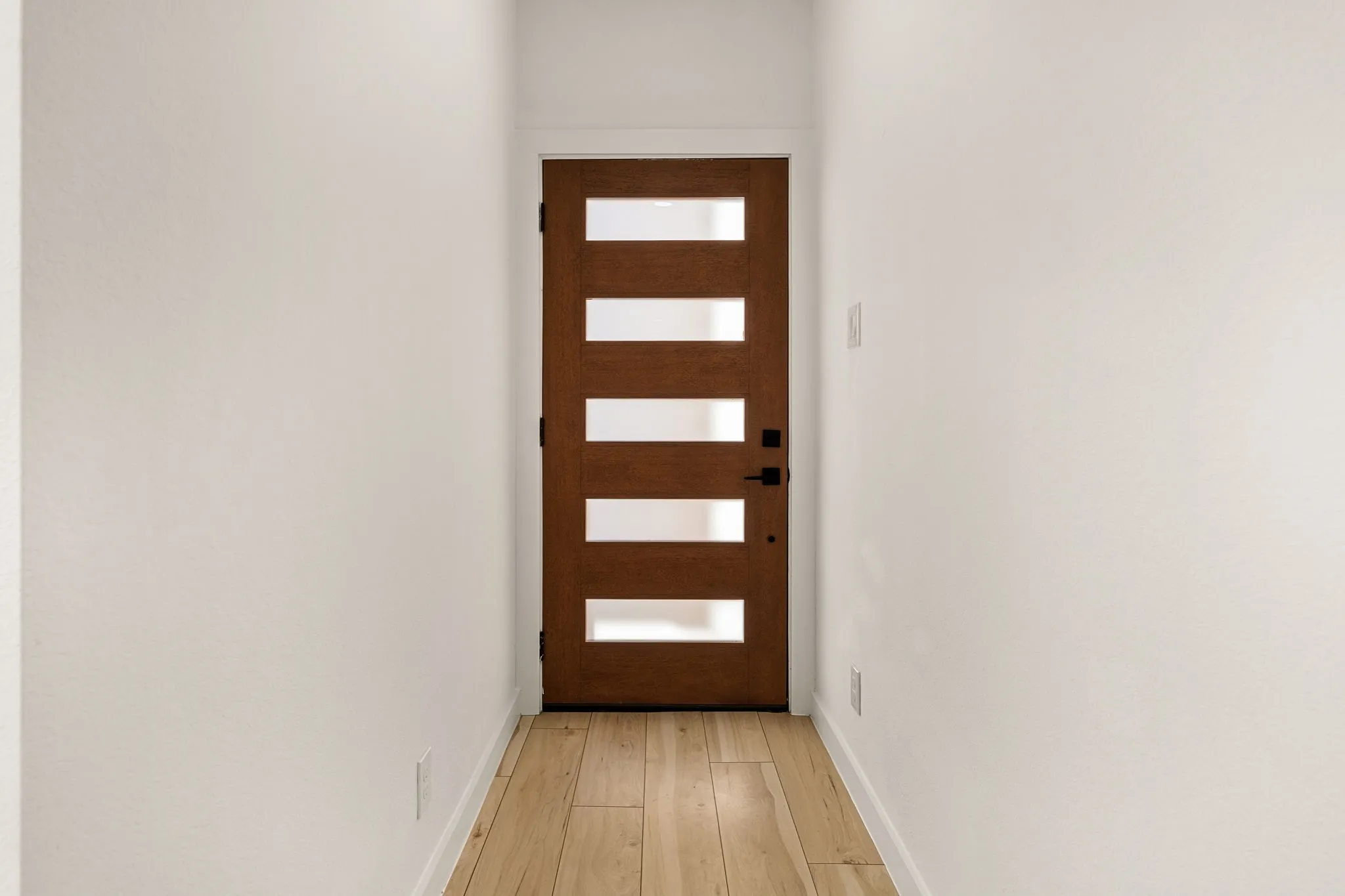Warm wood-tone entry door with frosted glass inserts, matte black hardware, and light wood-finish flooring