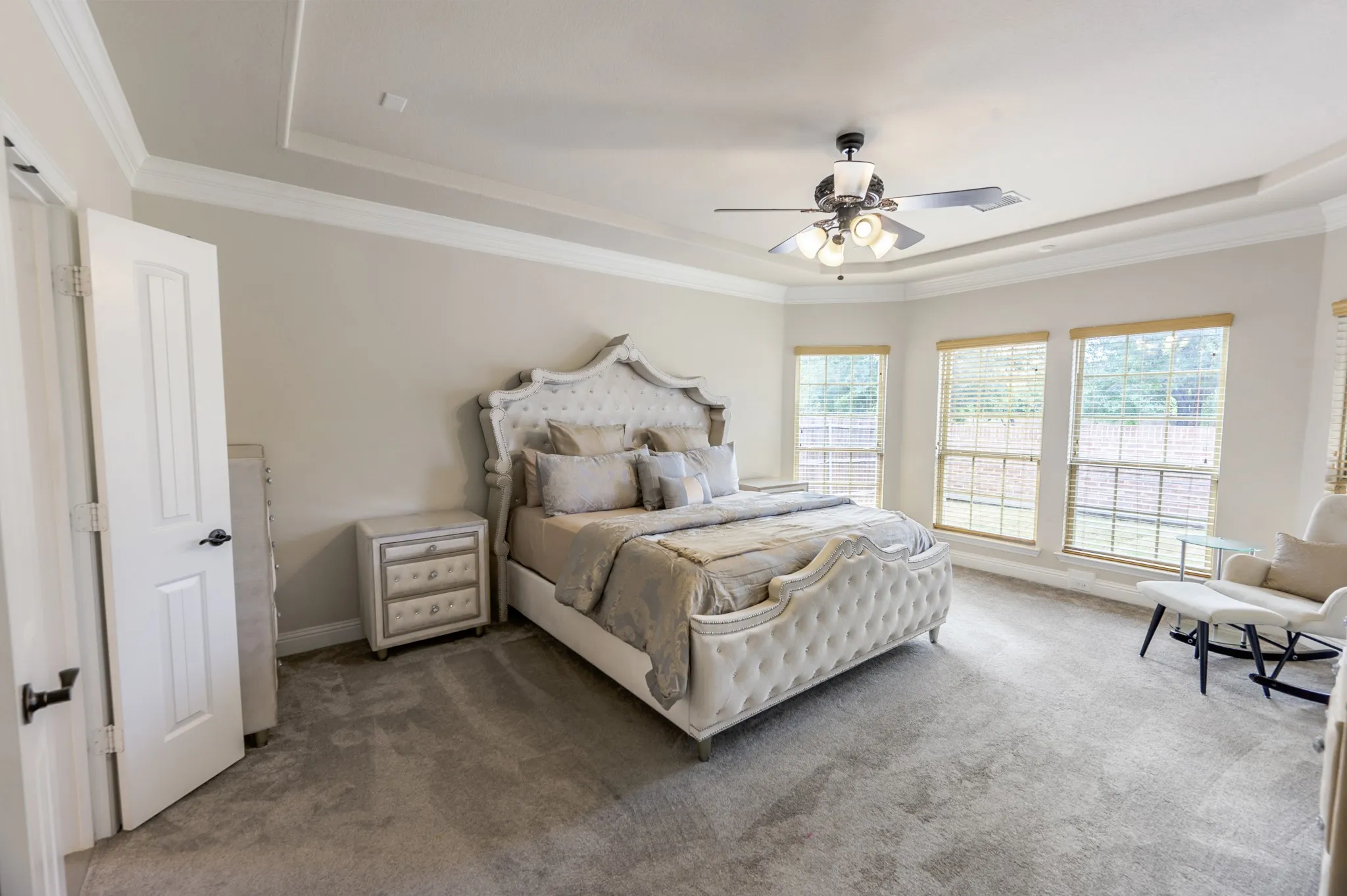 Tray ceiling with crown molding, ceiling fan with integrated lighting, three large windows, light gray wall paint, and gray carpet flooring