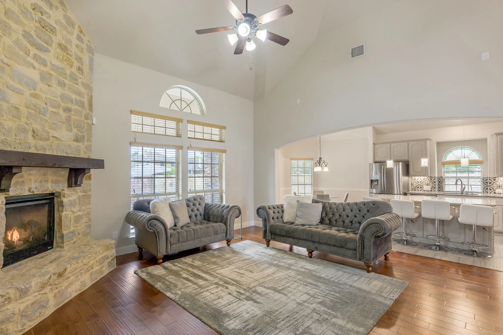 Vaulted ceiling, stone fireplace with wood mantel, wood-finish flooring, arched doorways, and a kitchen with an island featuring a breakfast bar