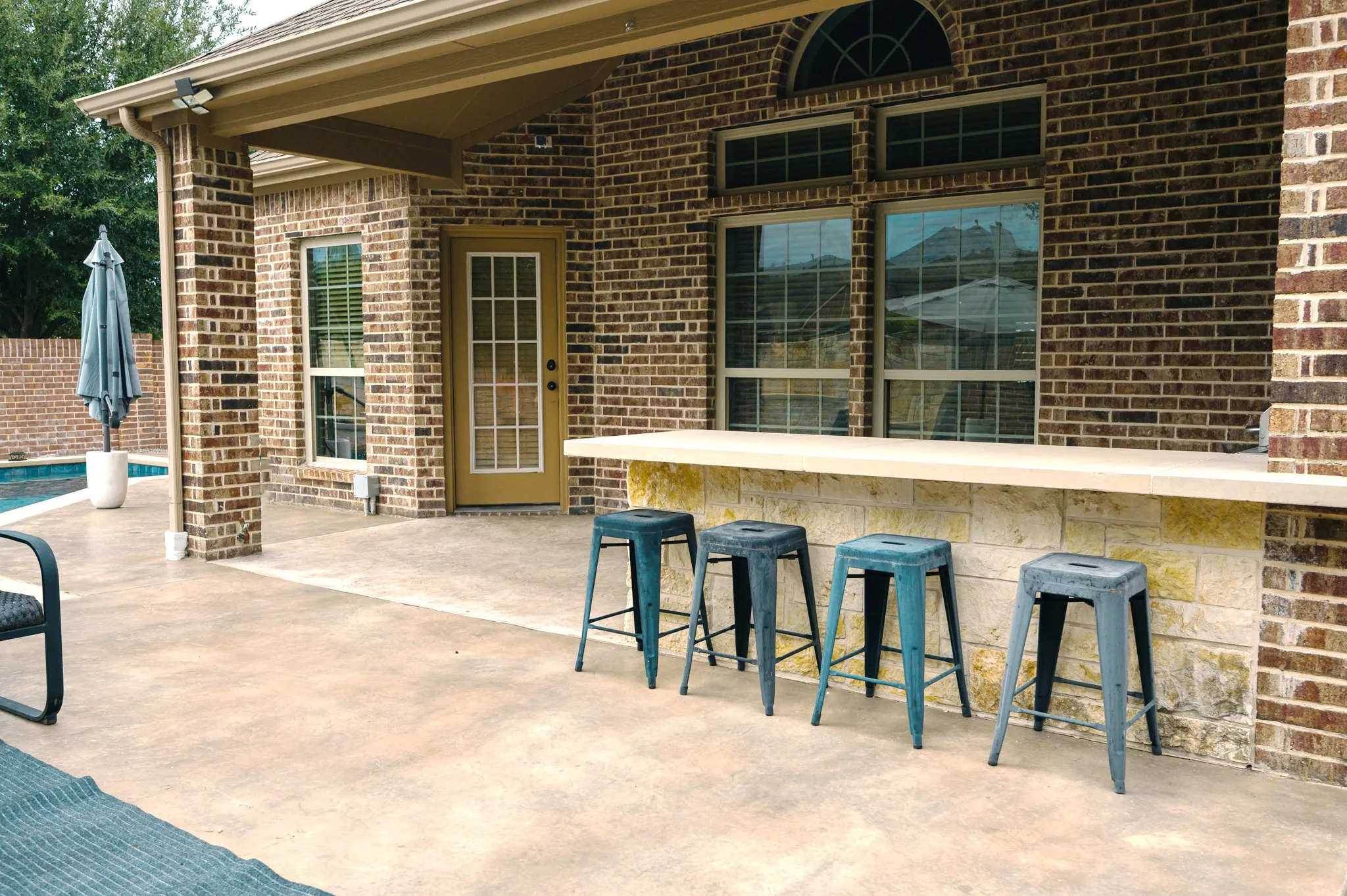 Brick exterior with a covered patio, featuring a built-in outdoor bar with a stone base and light-toned countertop, and a visible pool