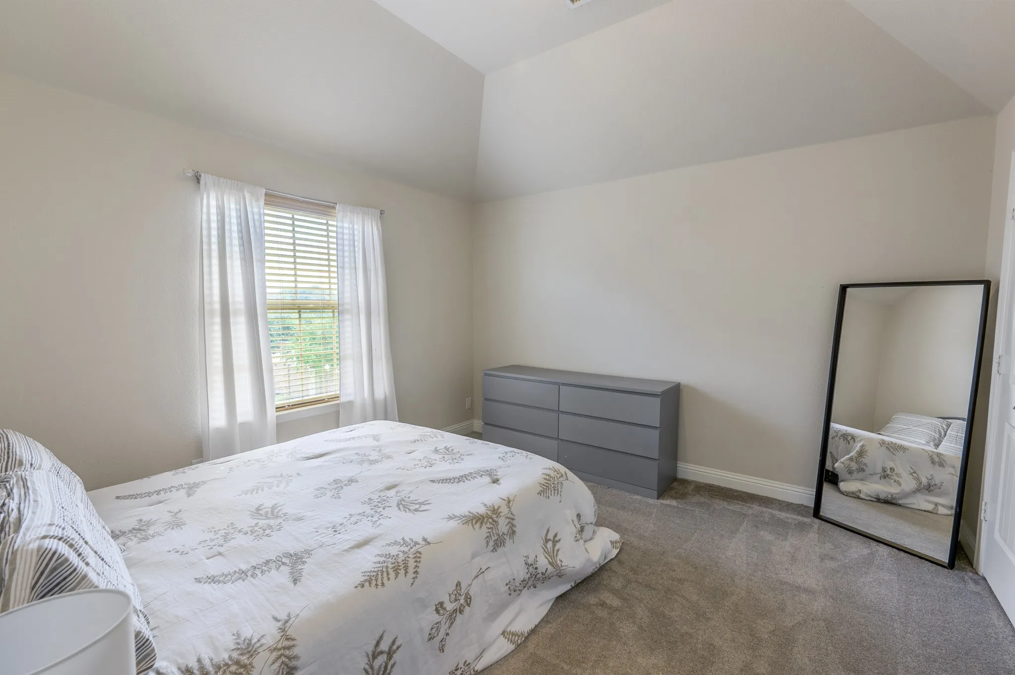Vaulted ceiling, light neutral wall color, window with blinds, carpeted flooring, and white trim