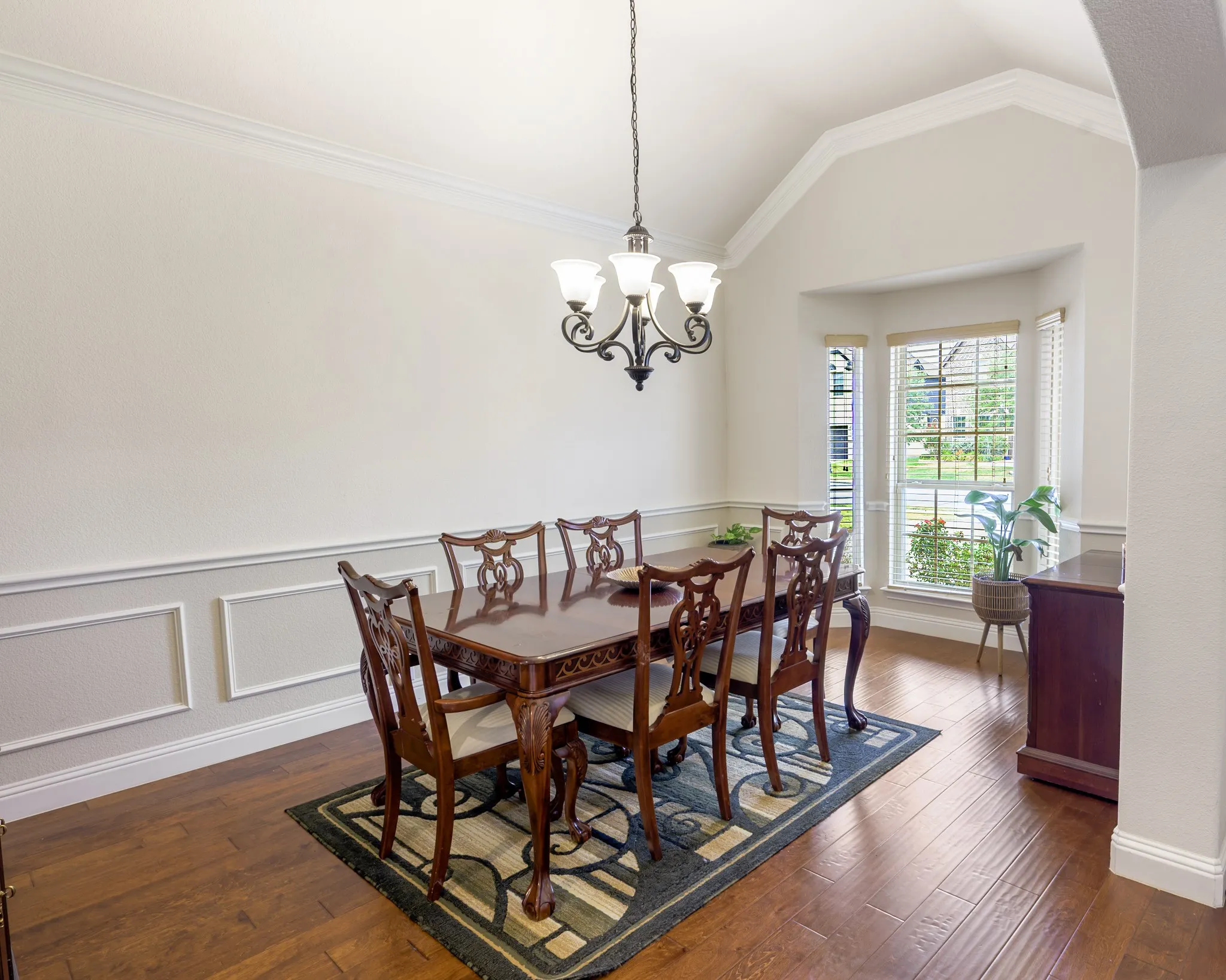 Angled ceiling with crown molding, wood-finish flooring, bay window, wainscoting, and a multi-light chandelier