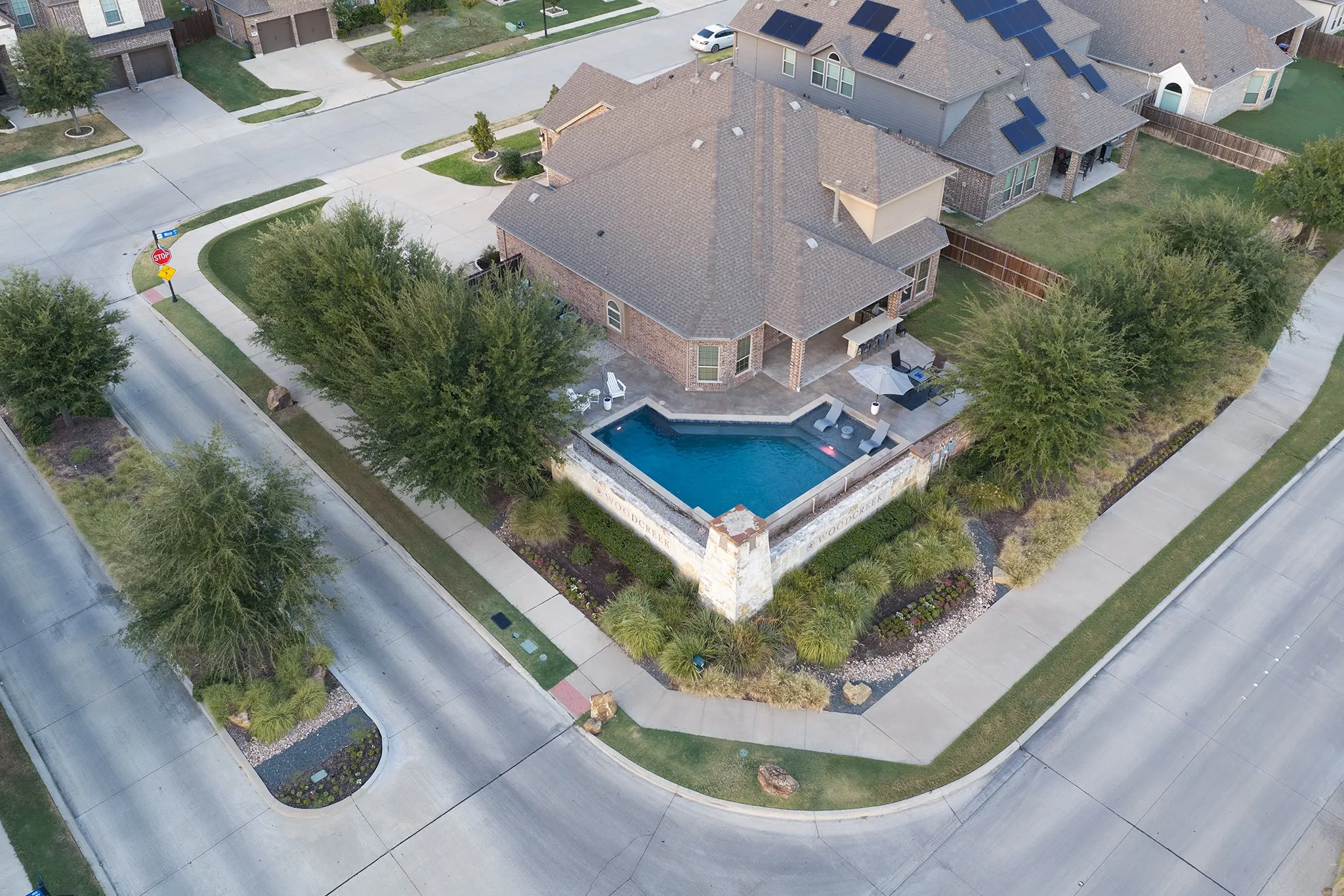 Corner lot residence featuring a geometric pool with stone surround, covered outdoor patio, and mature perimeter landscaping