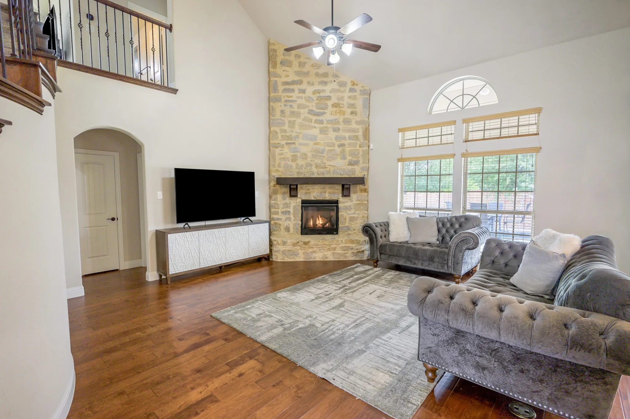 Vaulted ceiling, stone fireplace with a wood mantle, wood-finish flooring, two-story windows with an arched transom, and a wrought iron staircase railing