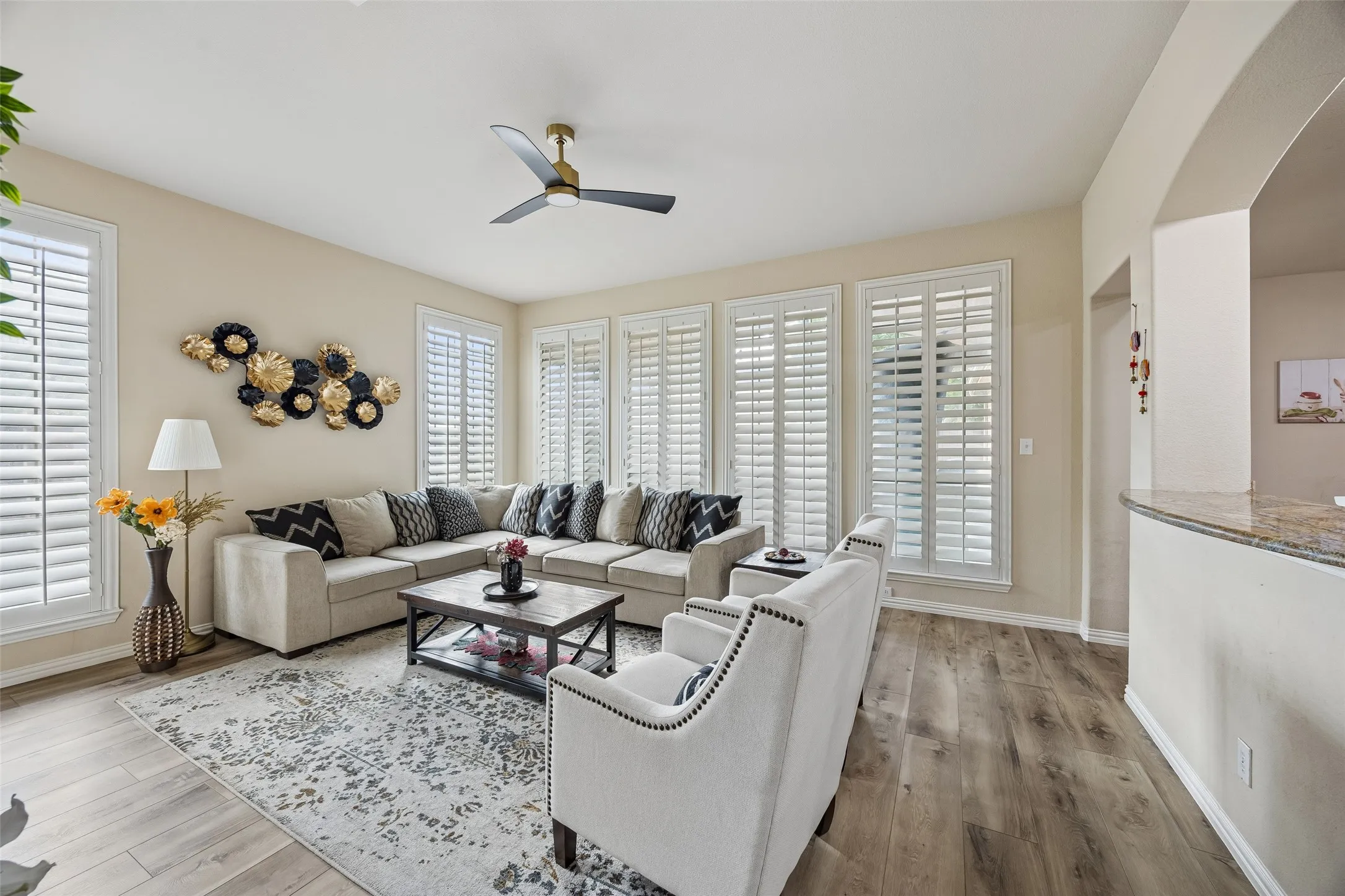 Spacious living area featuring wood-finish flooring, multiple windows with white plantation shutters, a ceiling fan, and an arched entryway