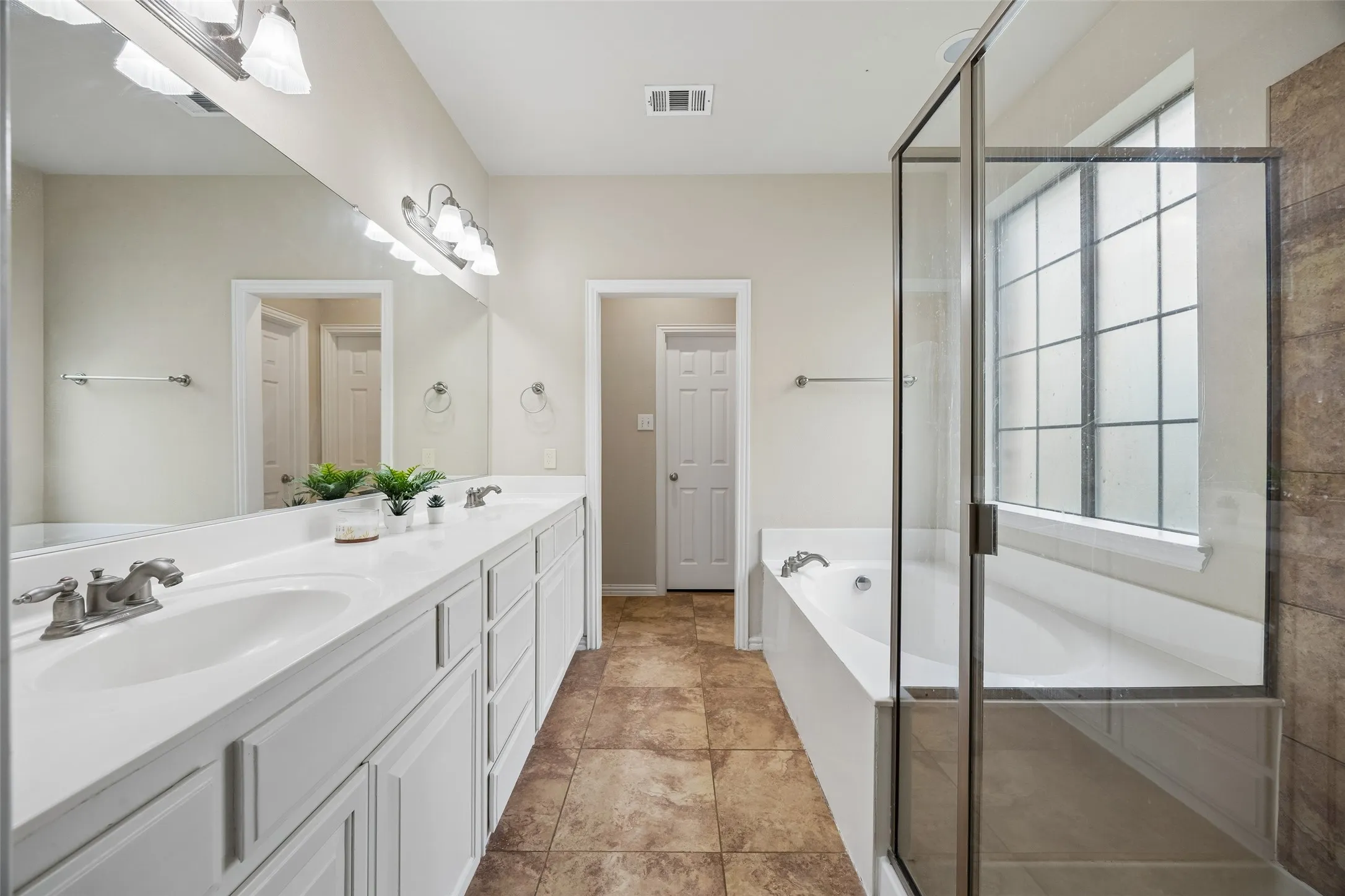Dual vanity with white cabinetry and integrated sinks, large wall-mounted mirror, separate soaking tub, and a glass-enclosed shower with tiled surround