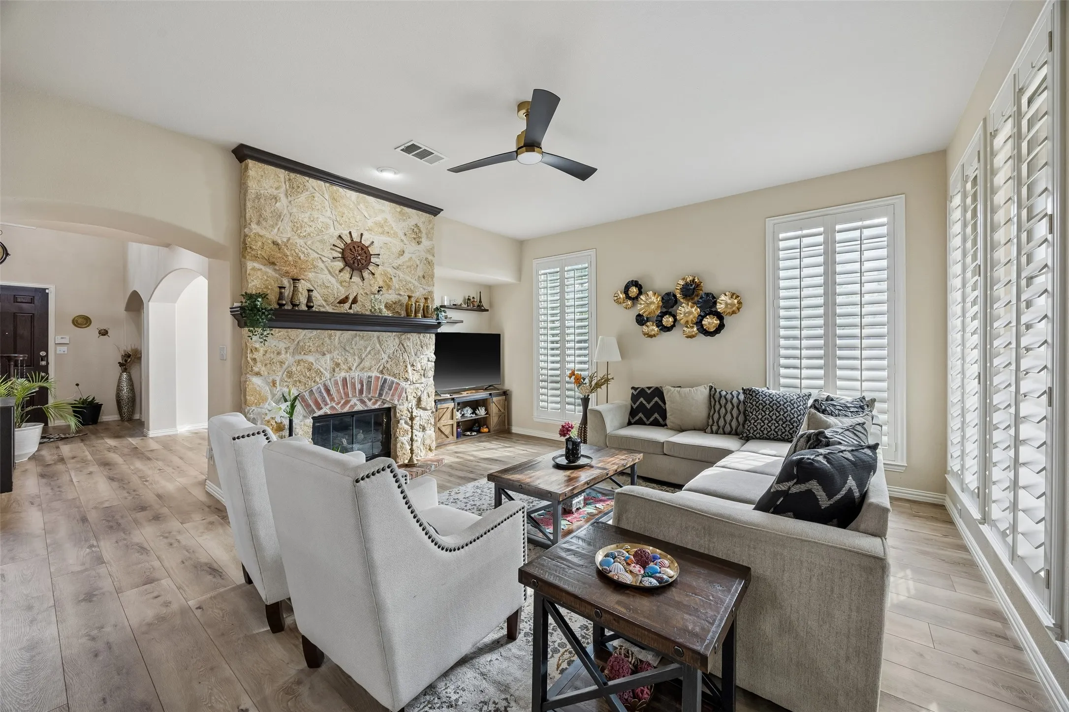 Stone-clad fireplace with a brick arch and dark wood mantel, wood-finish flooring, white plantation shutters, and a ceiling fan with integrated lighting