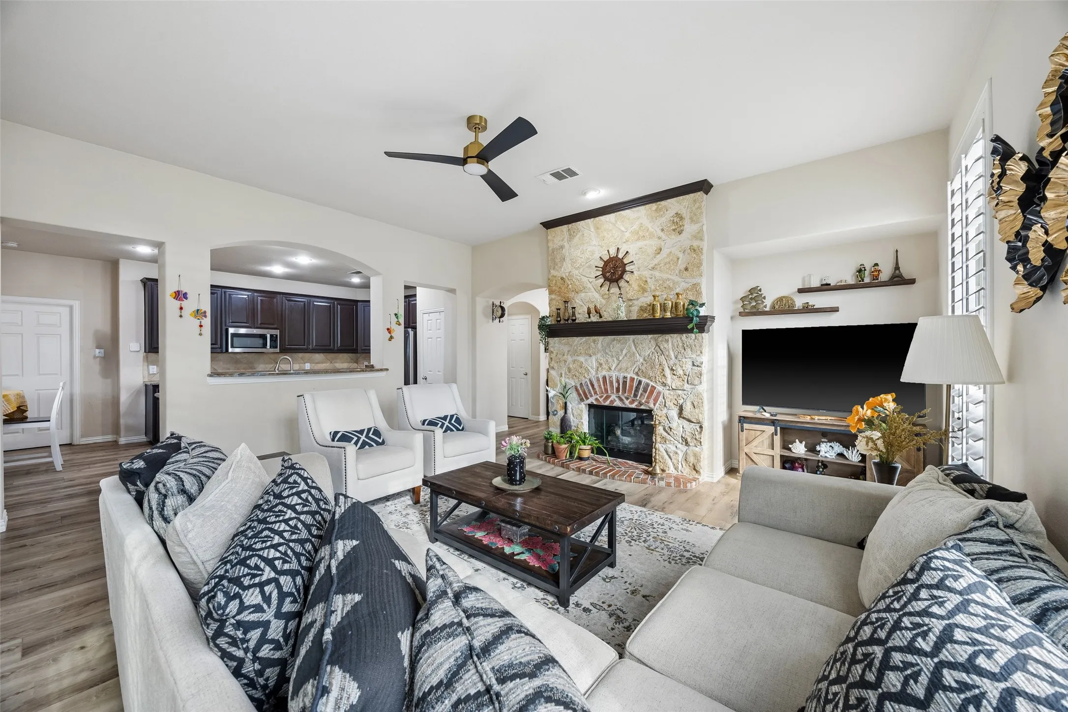 Stone-clad fireplace with brick interior and wood mantel, wood-finish flooring, ceiling fan, archway, and white plantation shutters