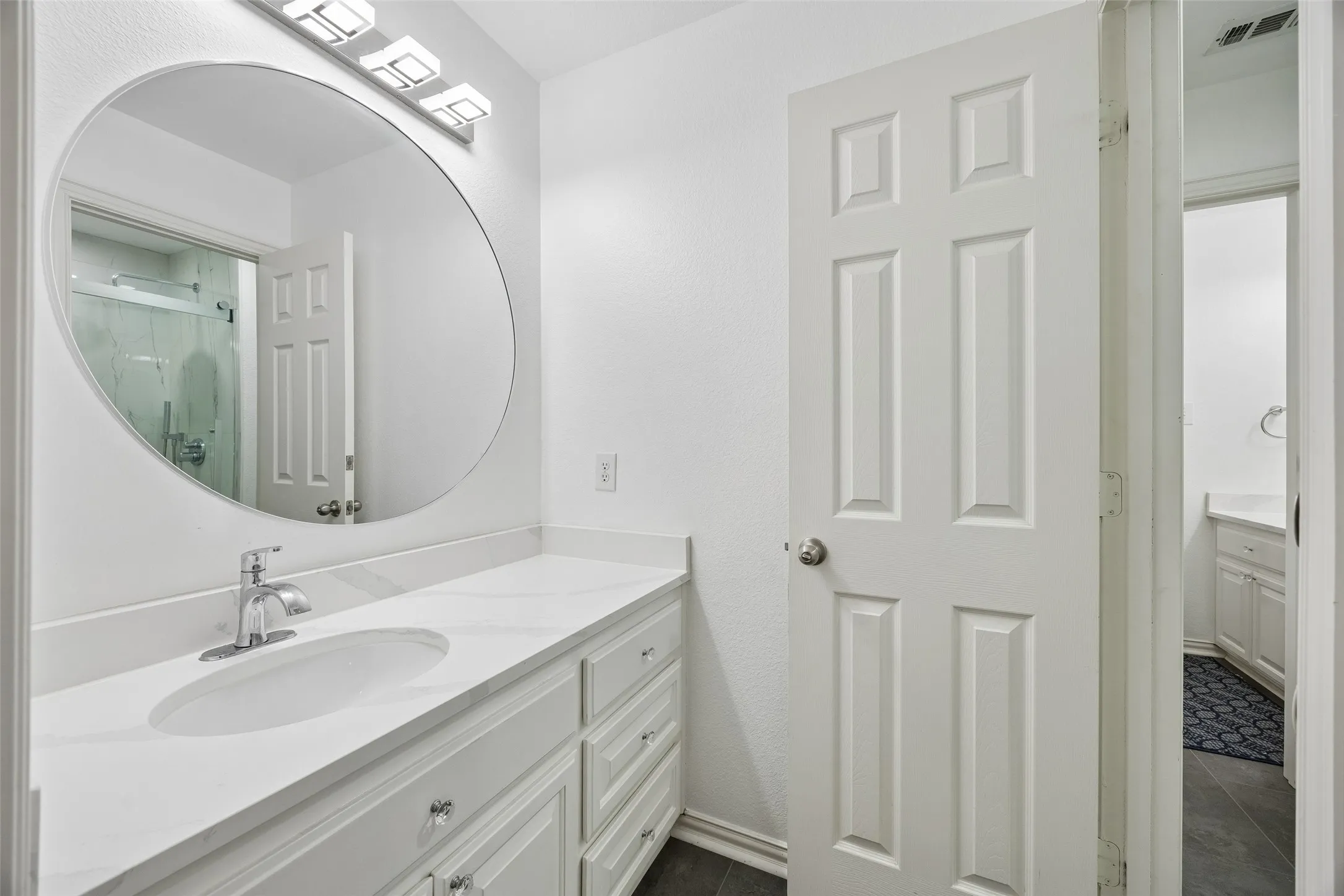 White paneled door opening to a vanity area with a chrome faucet, oval sink, and a circular mirror