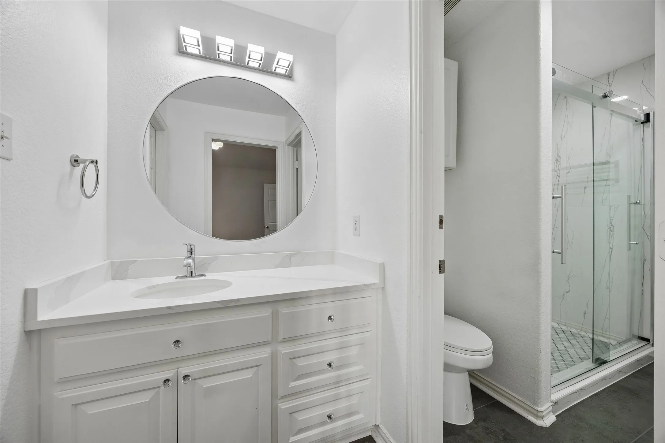 White vanity with integrated sink and chrome faucet, large circular mirror, four-light vanity fixture, glass-enclosed shower with subway tile, and dark floor tiling