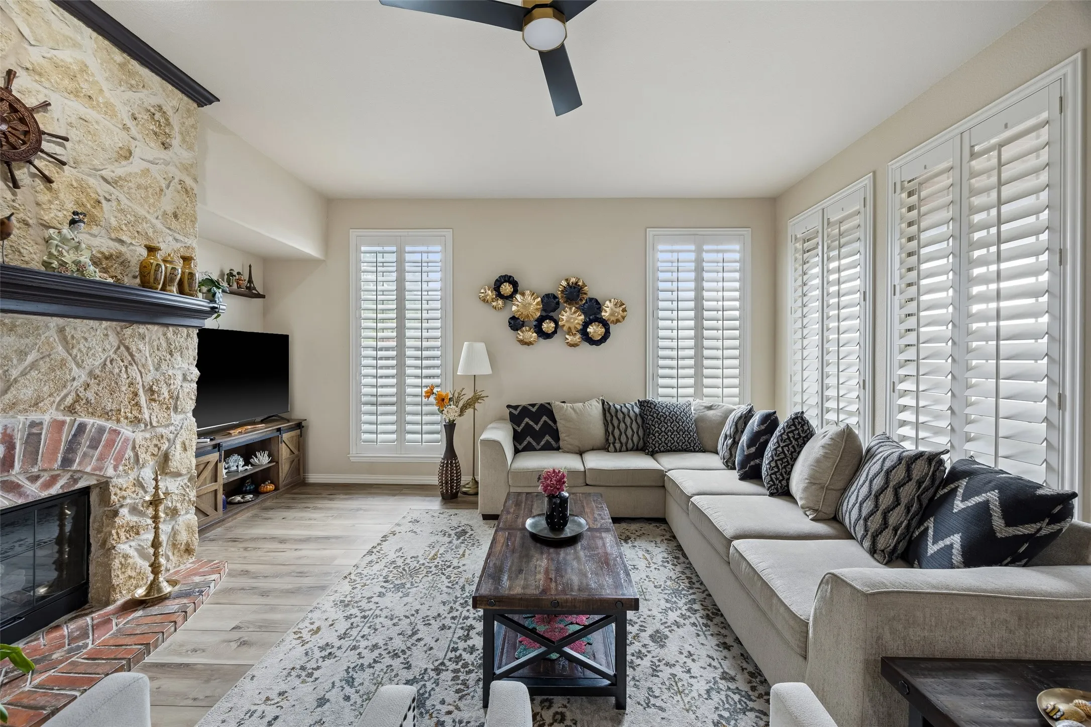Stone-clad fireplace with brick hearth, wood-finish flooring, plantation shutters, and a ceiling fan