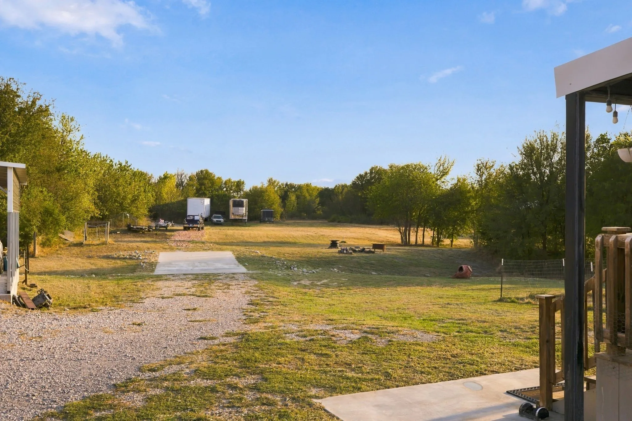 Expansive outdoor area featuring a gravel driveway, concrete pad, large grassy field, and mature trees